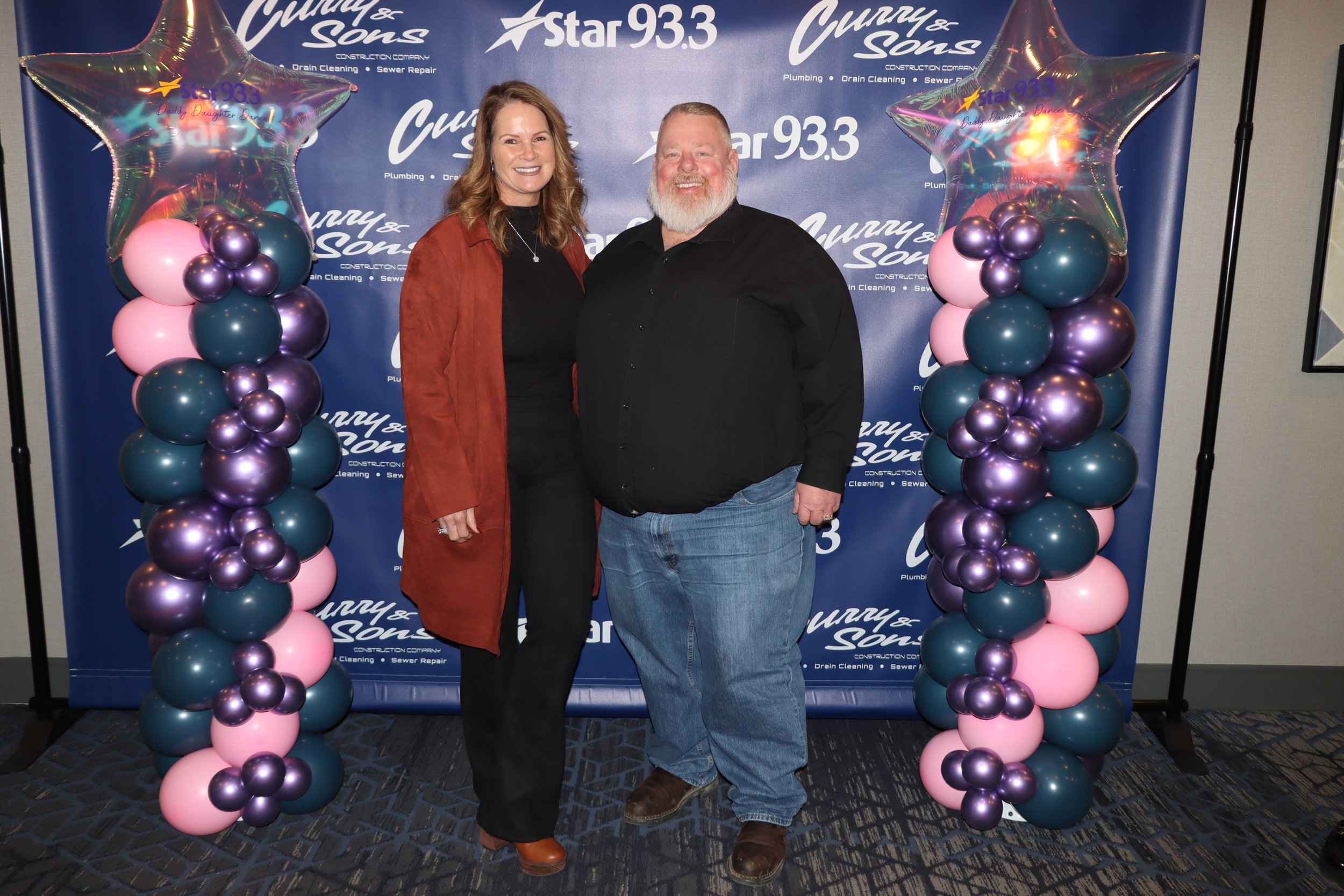 A woman and a man standing in front of a blue backdrop with the logos of 'Curry & Sons' and 'Star 93.3'. The backdrop is decorated with balloon pillars on each side, featuring pink, purple, and teal balloons topped with star-shaped balloons.