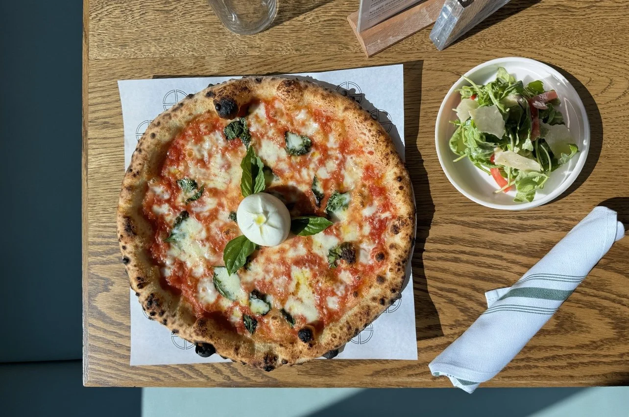 A pizza with cheese, basil, and tomato sauce on a wooden table, accompanied by a small bowl of salad and a rolled napkin.