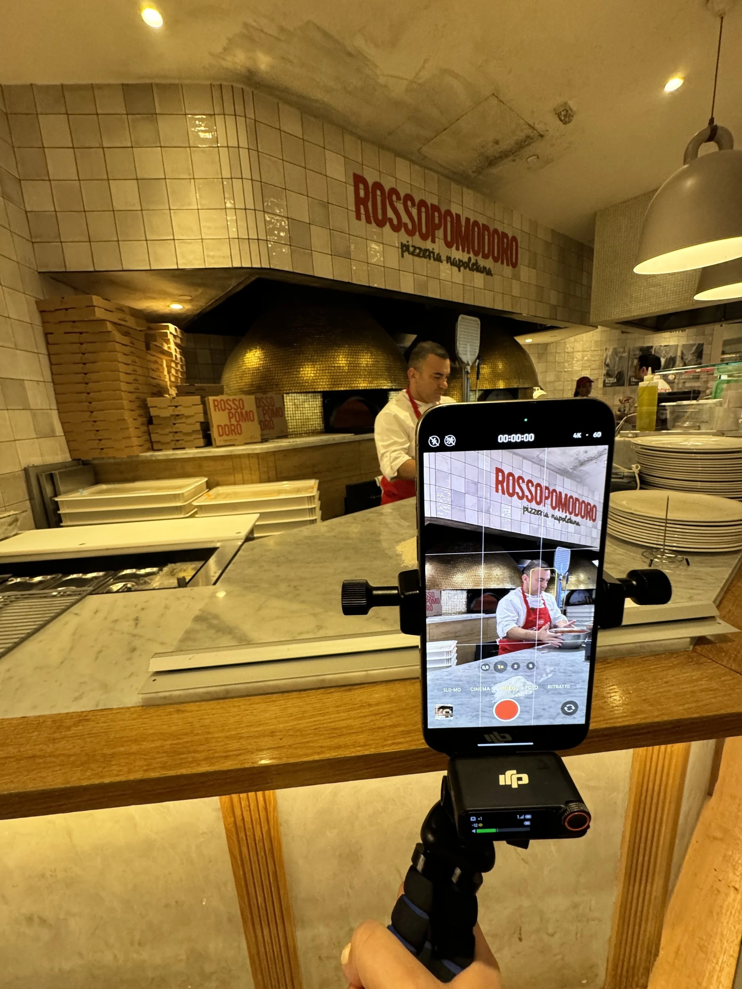 Photo of a person filming the interior of a pizza restaurant with a smartphone mounted on a stabilizer. In the background, a chef wearing a red apron and a white shirt is preparing food in front of a brick oven with a sign that reads 'ROSSOPOMODORO'.