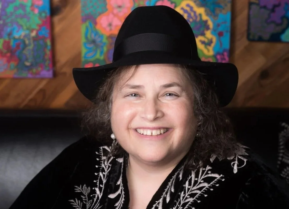A woman, artist Denise P. Levine, smiling, wearing a black hat and black clothing, sitting in front of colorful abstract paintings on a wooden wall.
