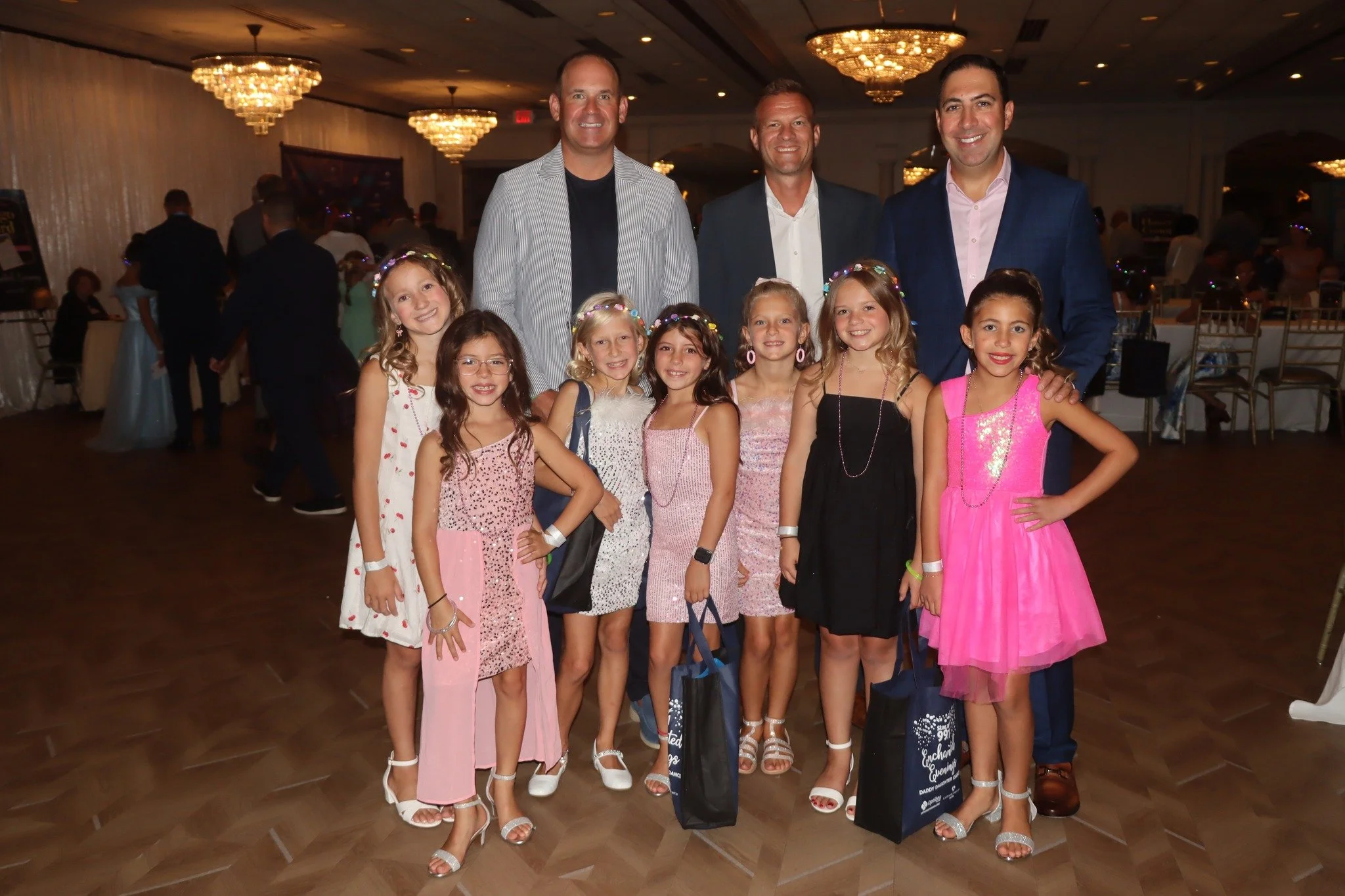 Group of nine young girls in dresses and three adult men in suits standing together at an indoor event, posing for a photo.