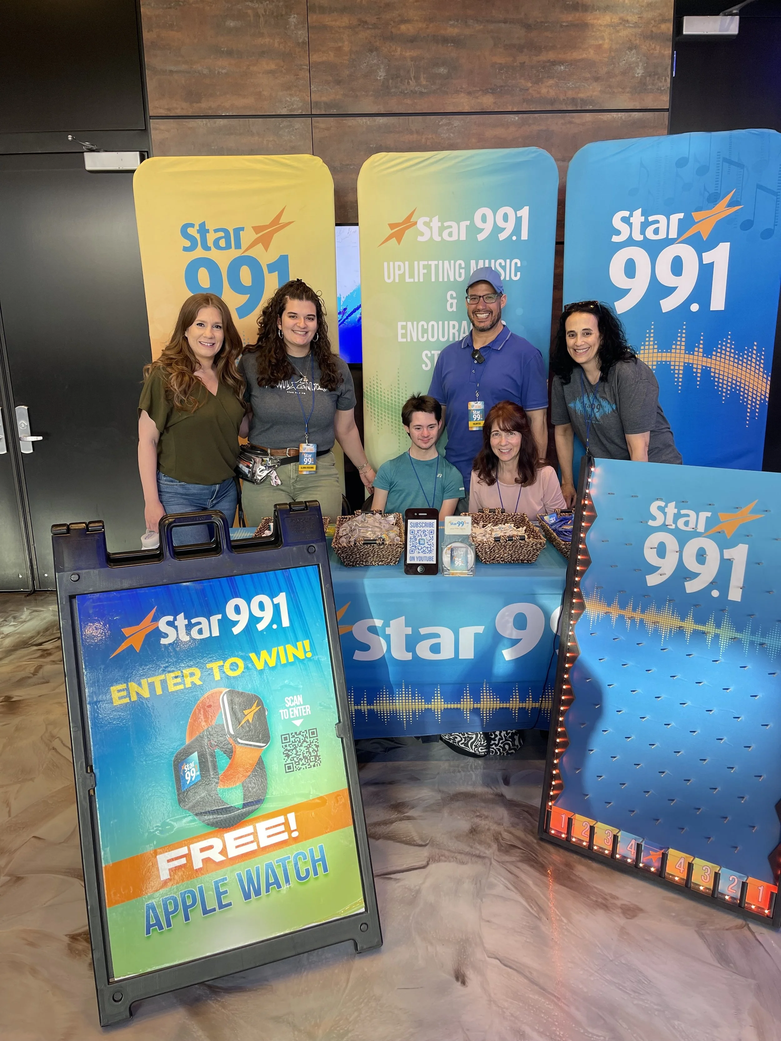 A group of six people at a promotional event for Star 99.1 radio station, standing behind a table with giveaway items, banners, and a contest sign indicating a chance to win a free Apple Watch.