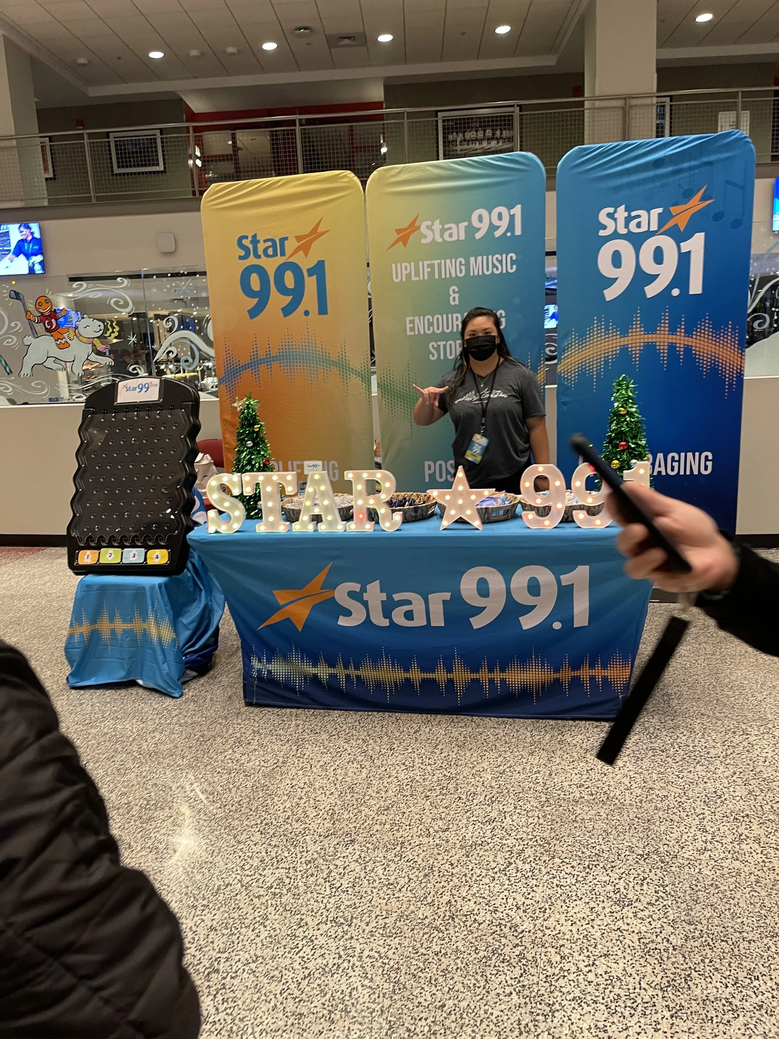 Radio station promotional booth for Star 99.1 with a table covered in a blue cloth displaying the station's logo, a woman in a gray shirt and black mask standing behind the table, decorative illuminated letters spelling 'STAR 99', small Christmas tre