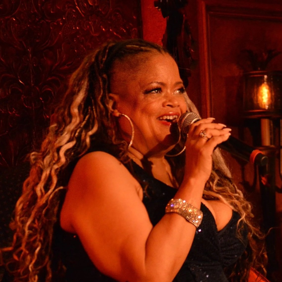 A woman with long, dreadlocked hair singing or speaking into a microphone on stage with warm lighting and wooden decor in the background.