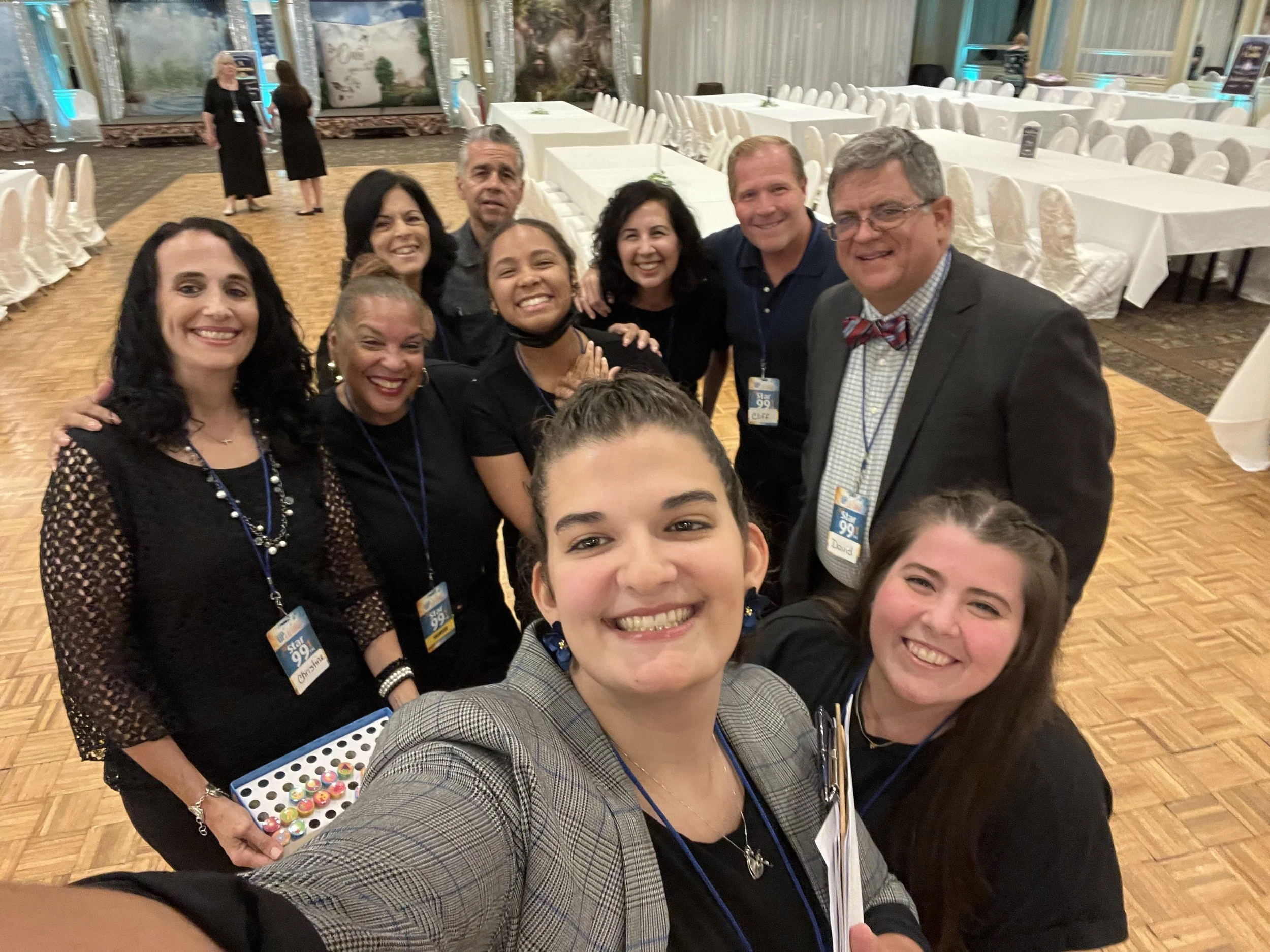 Group of nine smiling people taking a selfie at an indoor event, with round tables and chairs in the background.