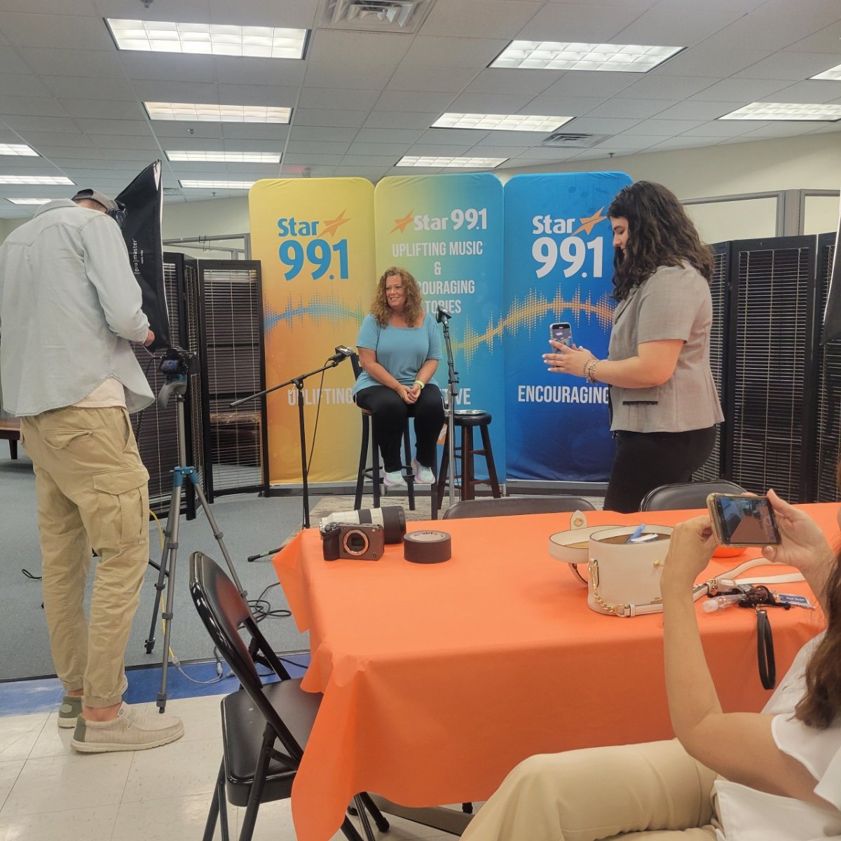 A woman sitting on a stool, being interviewed or recorded at a Star 99.1 event with a backdrop displaying the station's logo and slogan. A woman in the foreground is using a phone, and a person wearing a light jacket is filming with a camera. There i