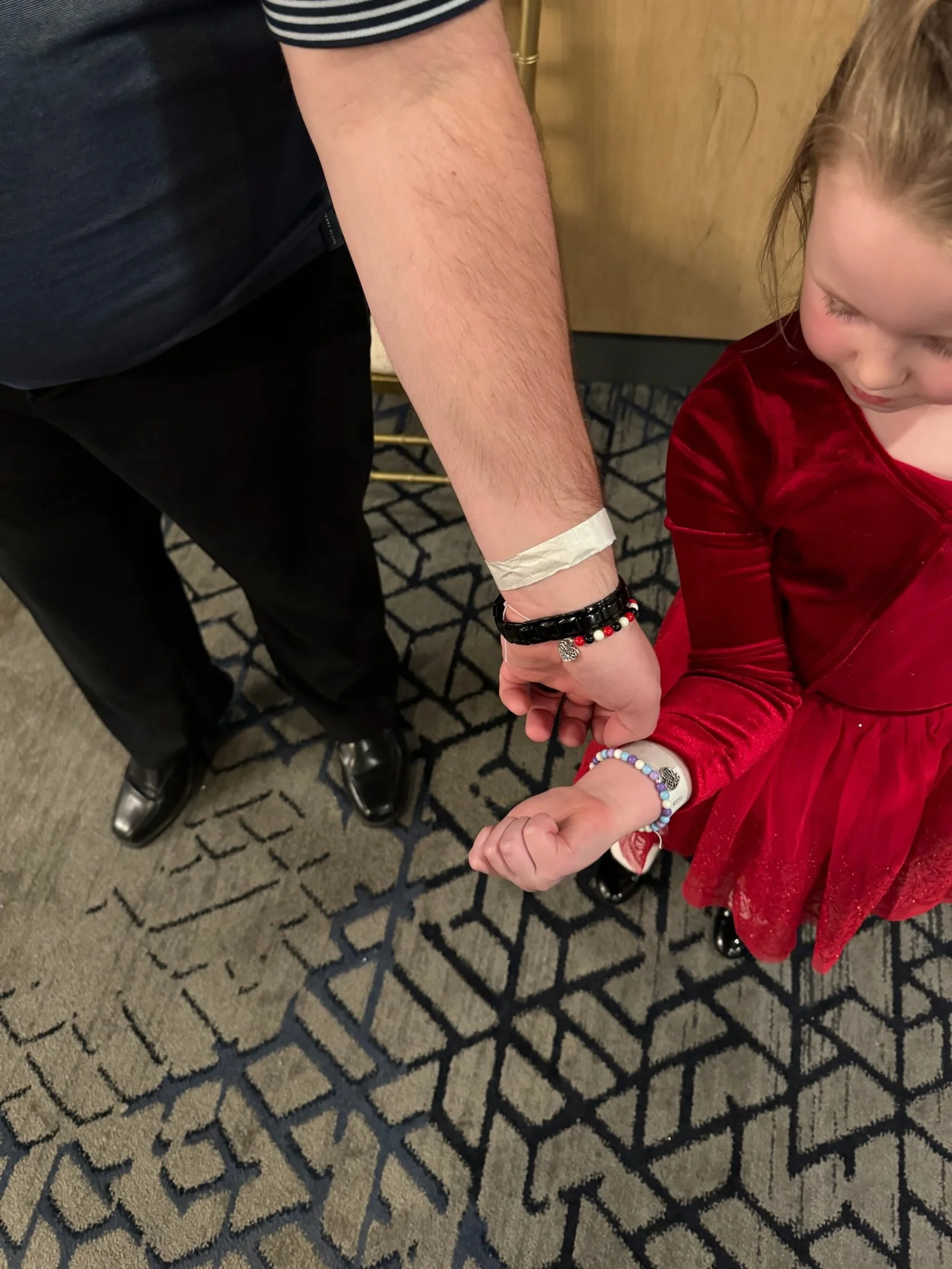 A man and young girl holding hands. The man is wearing black pants, a dark shirt with white stripes, and black shoes, with a medical wristband and beaded bracelet on his wrist. The girl wears a red velvet dress, colorful bracelets, and has a beaded b
