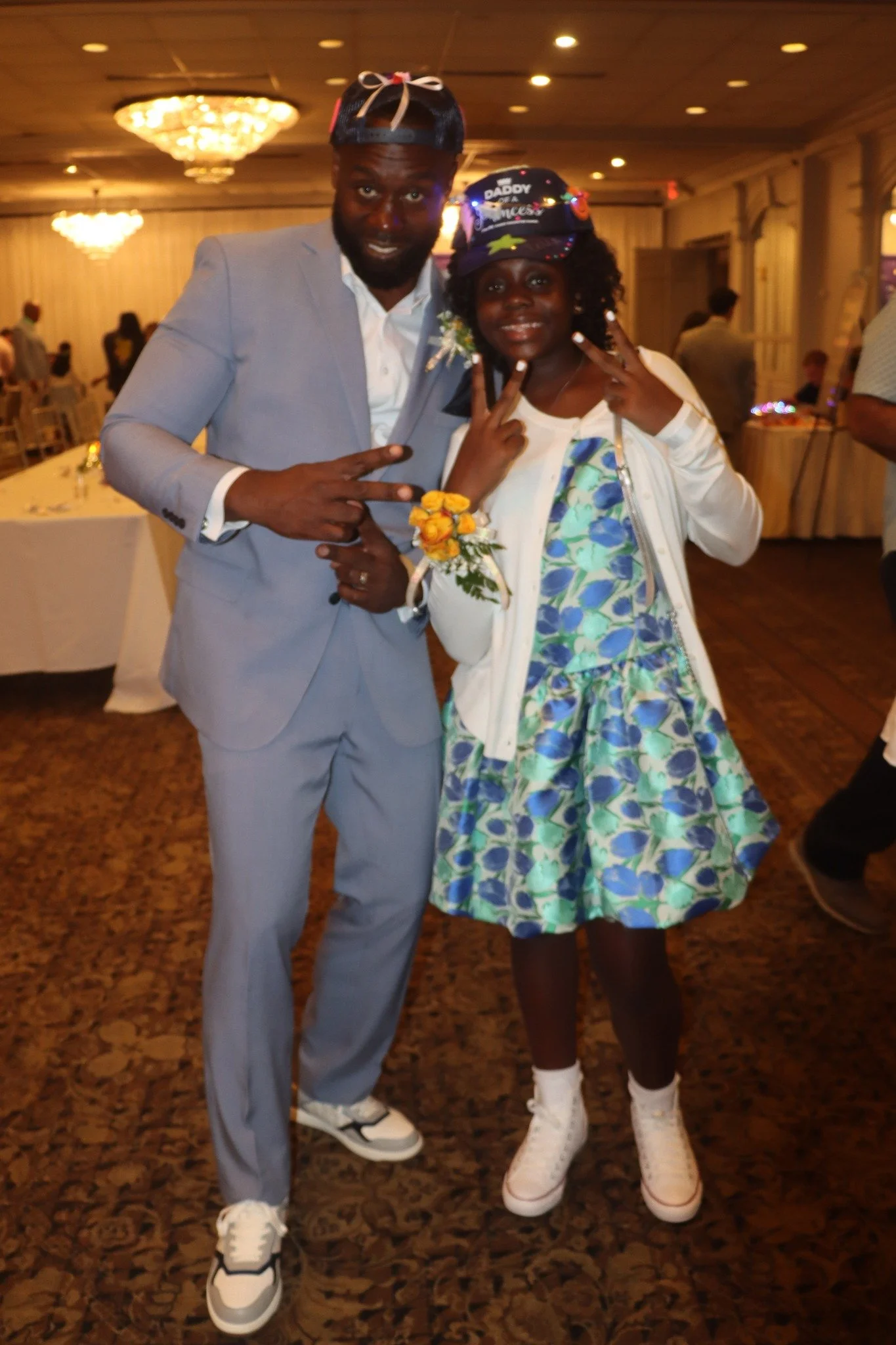 A man in a light gray suit and a girl in a colorful dress and jacket at a celebration, both making peace signs, with a decorated room in the background.