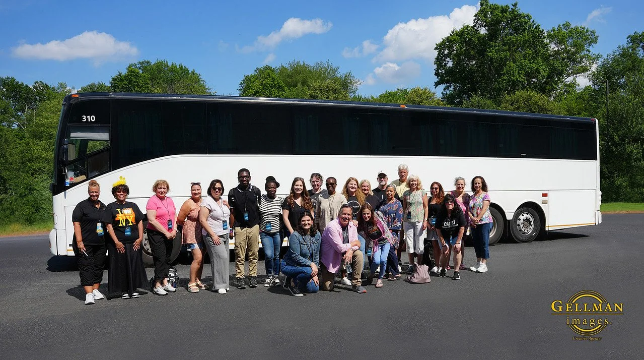 Group of diverse people standing in front of a large white tour bus on a paved road with green trees and a blue sky in the background.