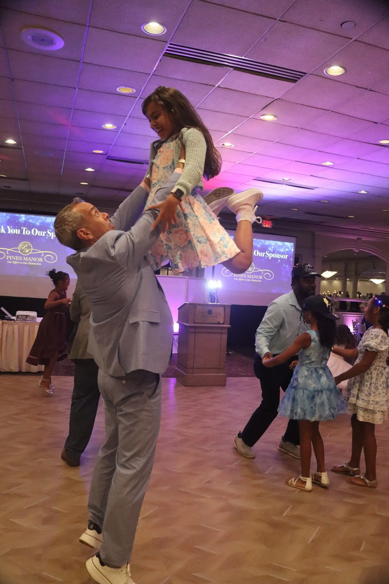 A man lifting a young girl, both smiling, at a dance reception with dancing guests and a decorated venue in the background.
