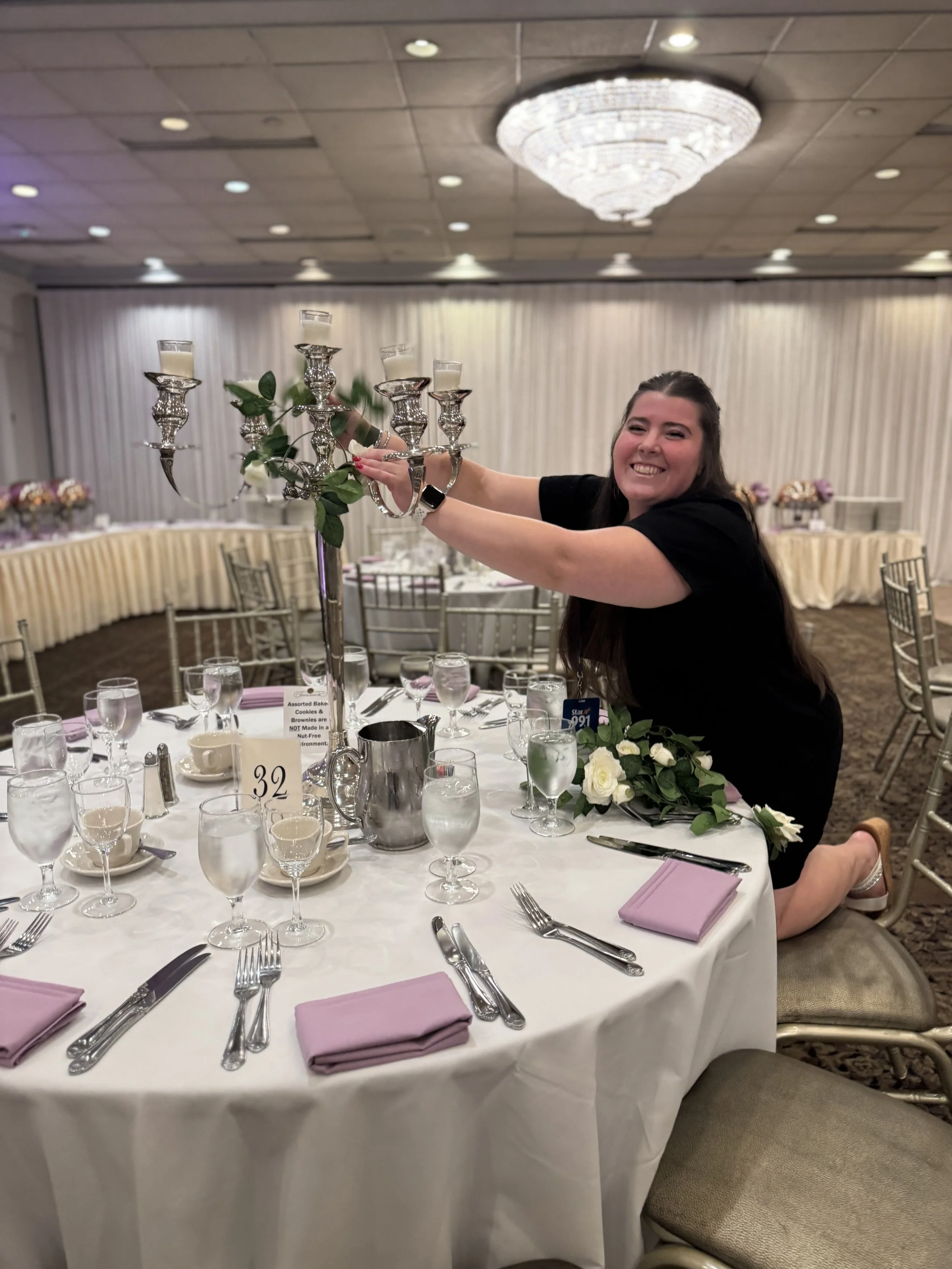 A woman kneeling on a banquet table, lighting a candelabrum with five cups, in a decorated event hall with fancy ceiling chandelier and round tables with place settings, pink napkins, and floral centerpieces.