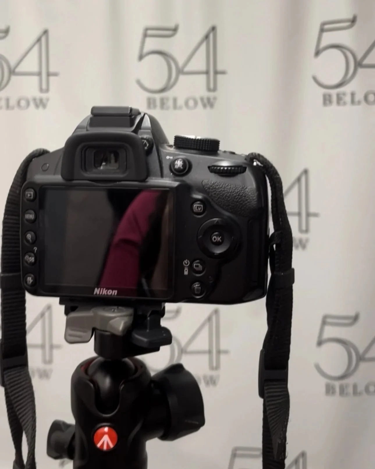 A Nikon DSLR camera mounted on a tripod, with a background showing the number 54 and the word 'Below' repeated in a pattern.