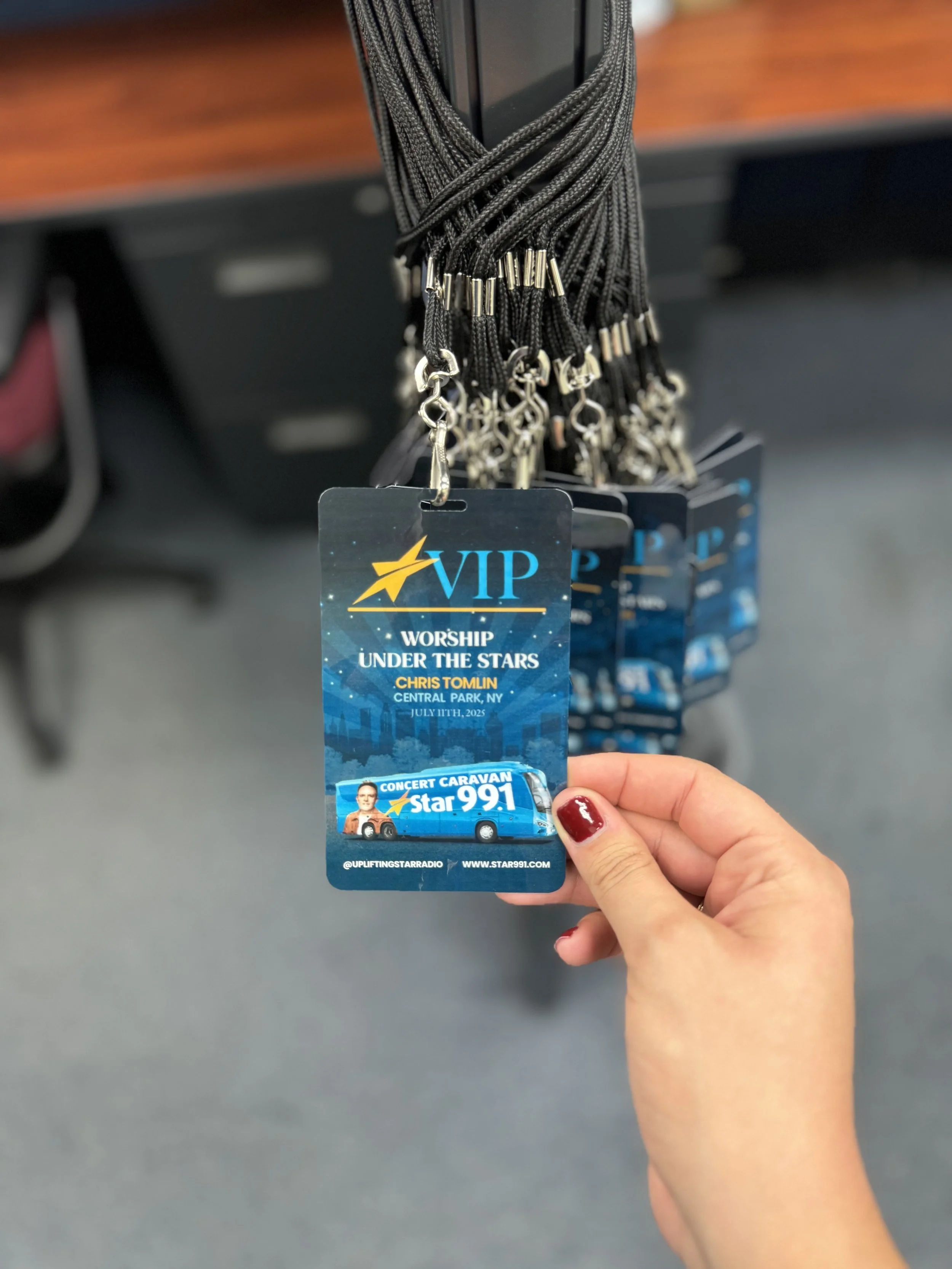 Hand holding a VIP concert ticket for worship under the stars event in Central Park, NY, featuring Star 99.1, with a background of additional similar tickets hanging from lanyards.