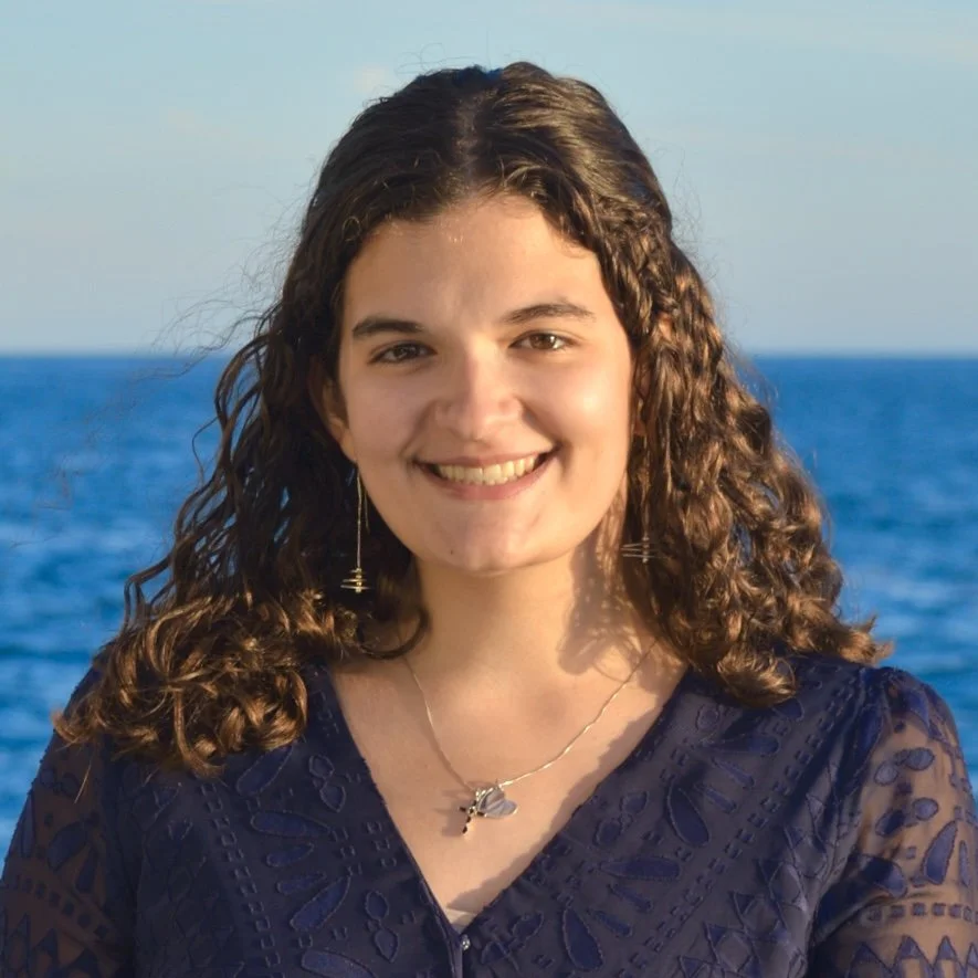 A young woman, Alanna Kroening, with curly brown hair smiling at the camera with the ocean in the background.