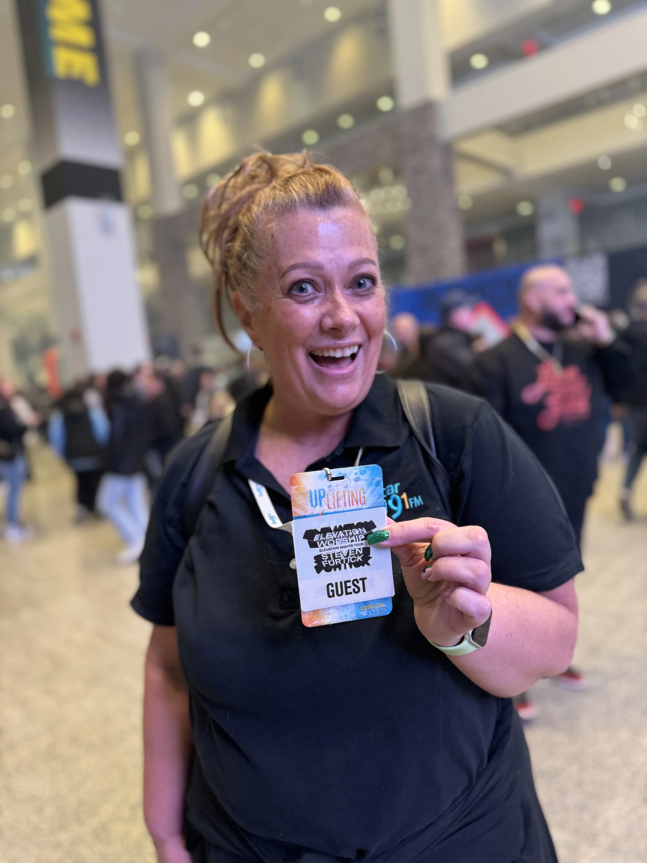 A smiling woman with red hair styled in dreadlocks, wearing a black shirt and a guest badge, holding it up at an indoor event with many people in the background.