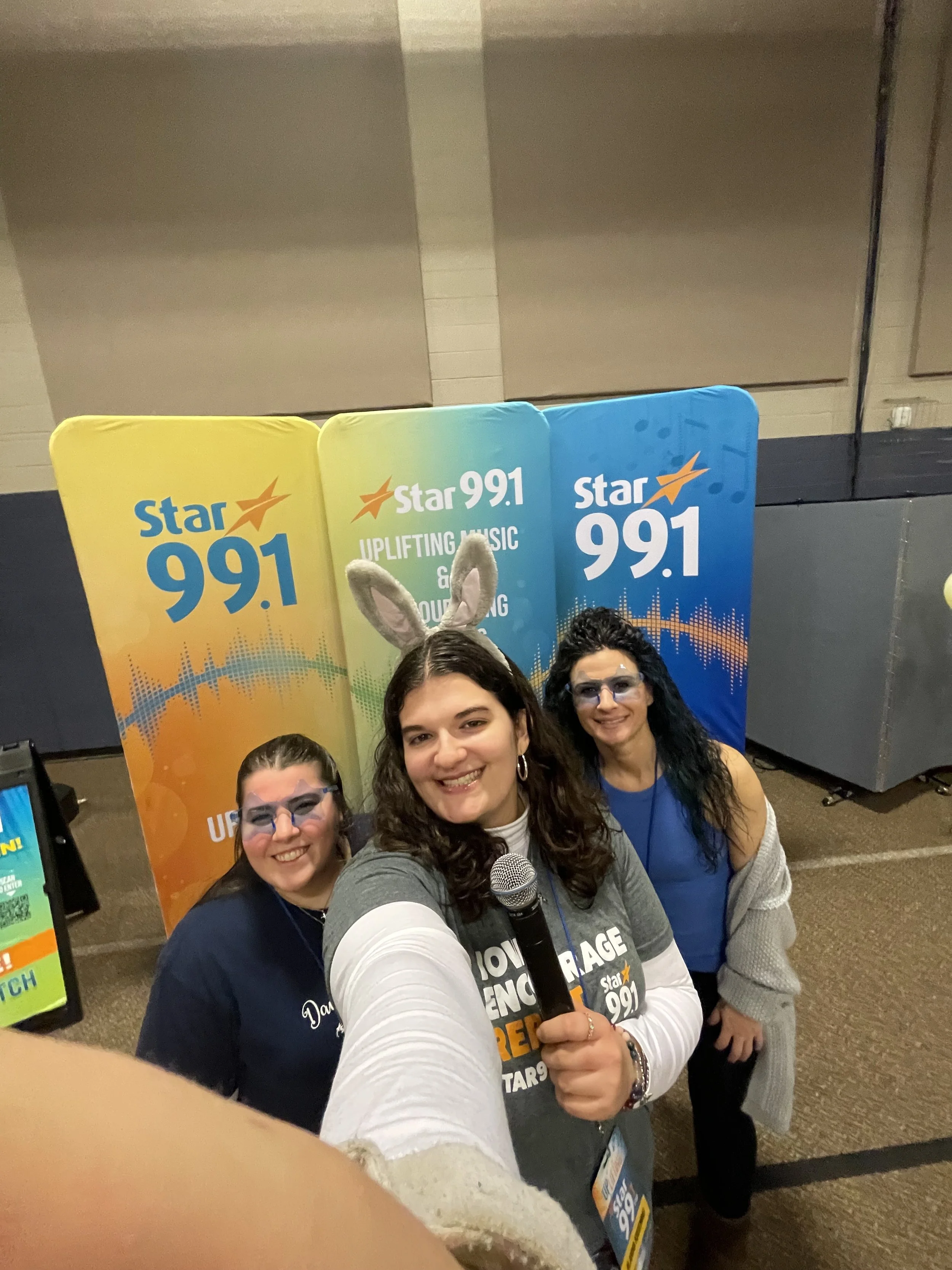 Three women at a promotional event for Star 99.1 radio station, posing for a selfie with a woman in the middle holding a microphone, one wearing bunny ears, and all wearing glasses with music-themed backgrounds in the background.