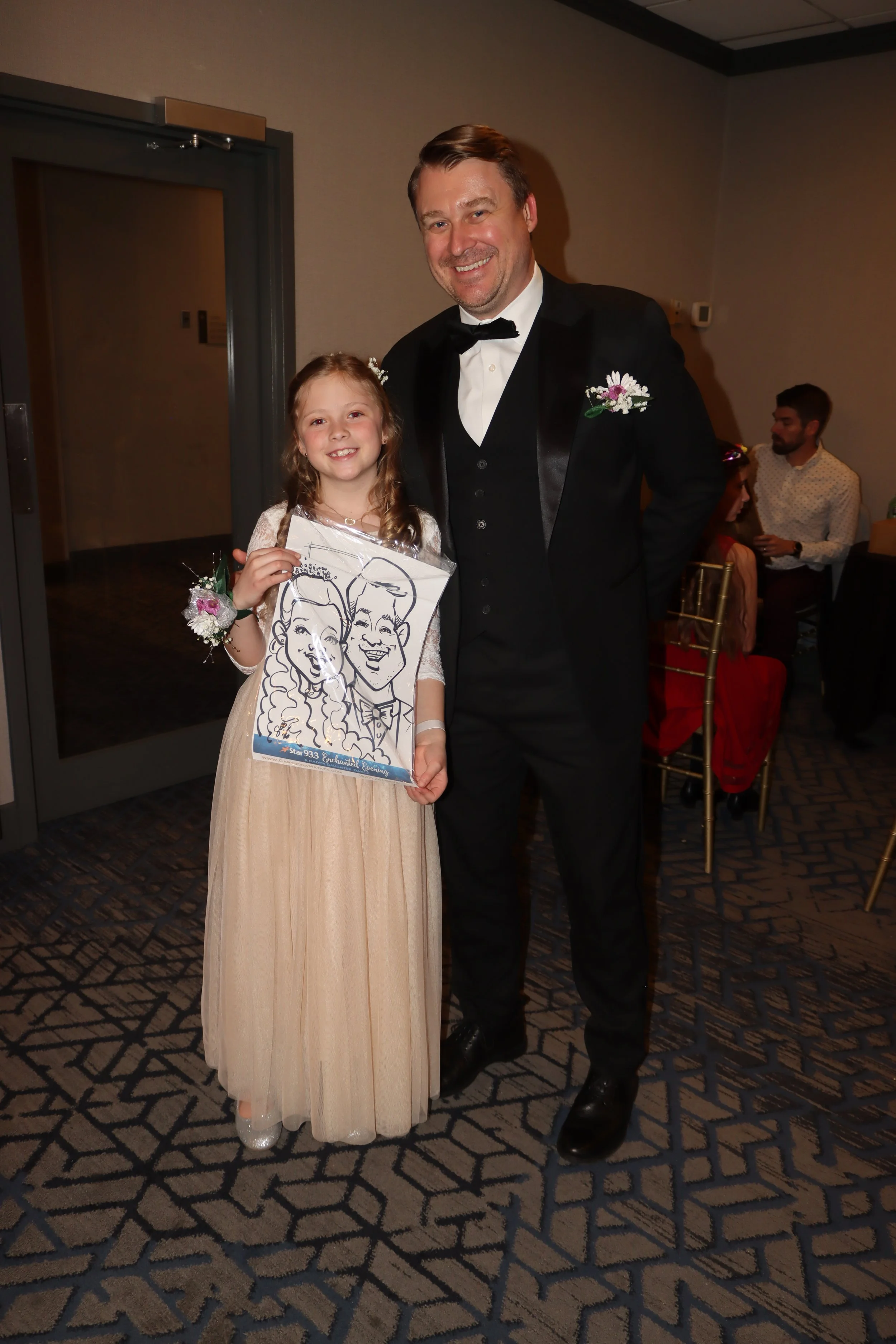 A young girl dressed in a beige gown holding a caricature of herself and a man in a tuxedo standing indoors at a wedding reception.