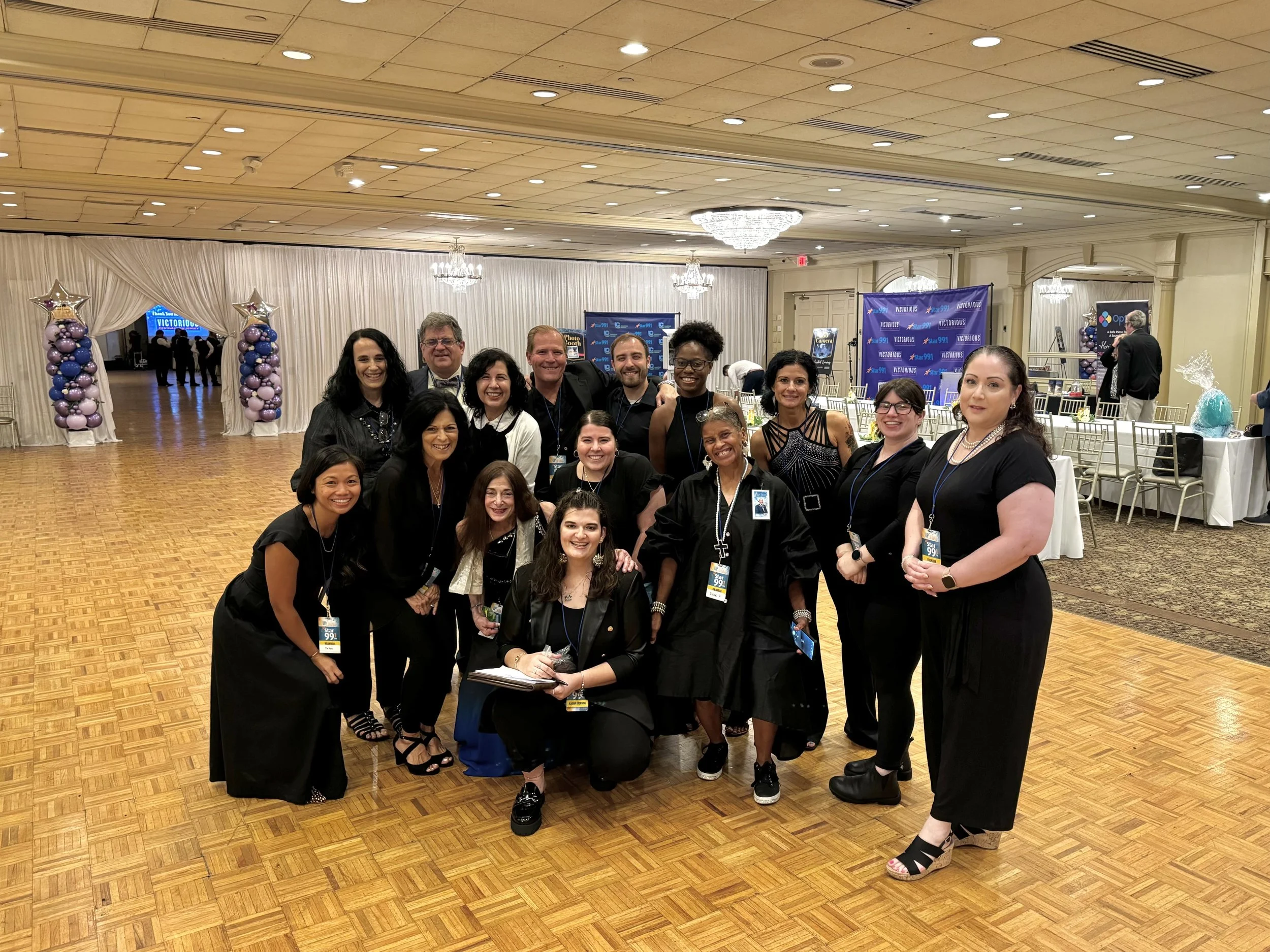 A group of 14 people posing for a photo in a banquet hall decorated with purple balloons and drapes, with tables and chairs in the background.