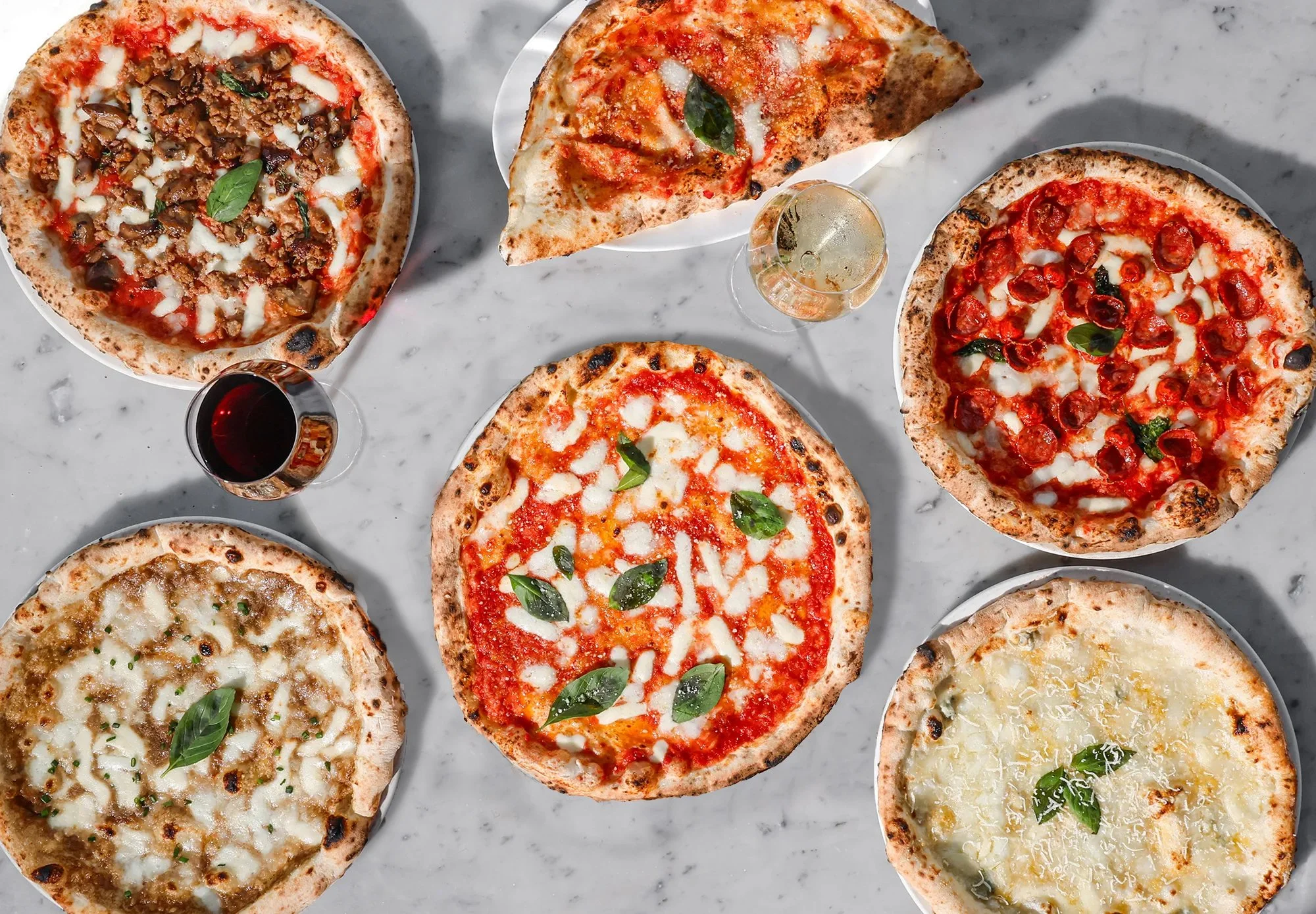 Six pizzas with various toppings on a marble table, along with glasses of red and white wine and a glass of sparkling wine.
