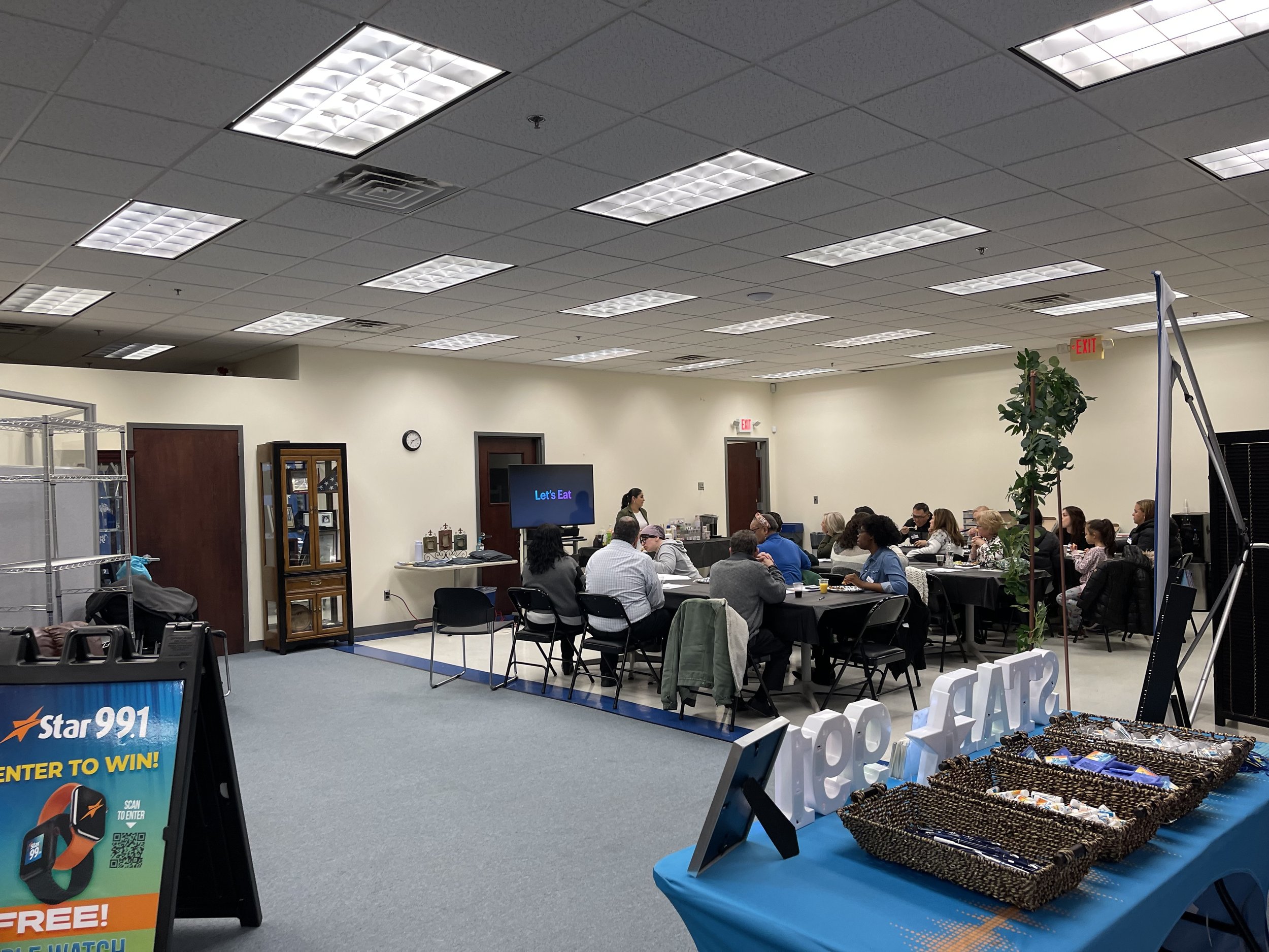 A conference room with people attending a presentation. There are tables with chairs, a large screen displaying 'Let's Eat,' and a sign for Star 99.1 radio station near the entrance. The room has a drop ceiling with fluorescent lights, a wall clock, 