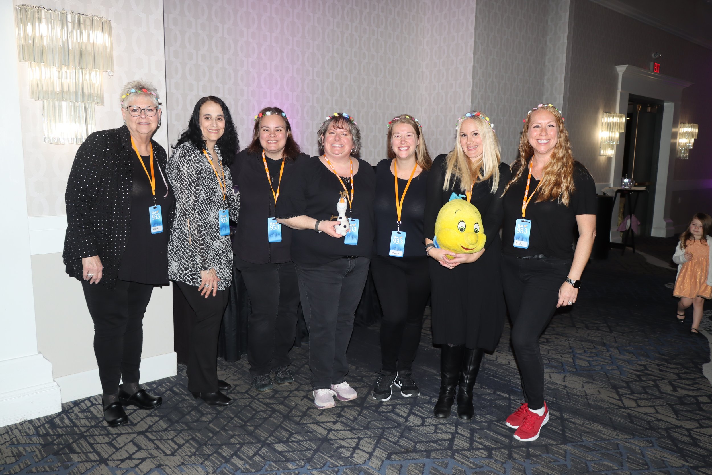 Group of eight women standing together in a hotel conference room, wearing black clothing and orange conference badges, some holding Halloween-themed plush toys, with two young children in the background.