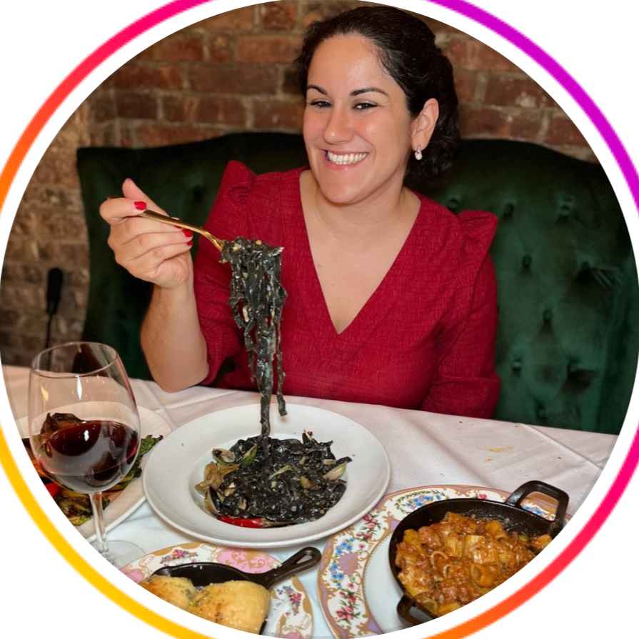 Woman in red dress smiling at table with black pasta, red wine, and other dishes.