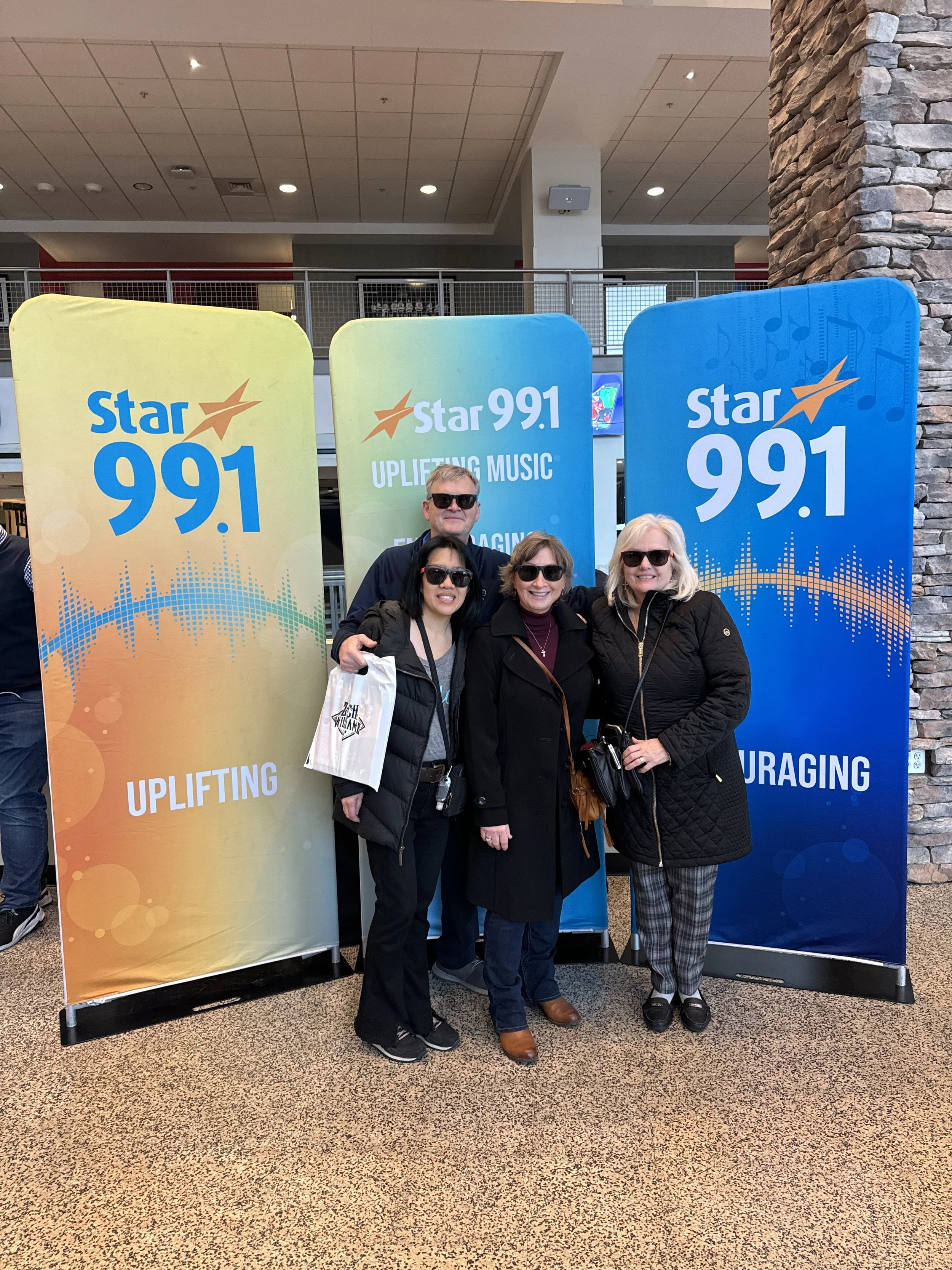 Four people standing together posing for a photo in front of three large banners for Star 99.1 radio station, inside a building with a stone wall and a first-floor balcony in the background.