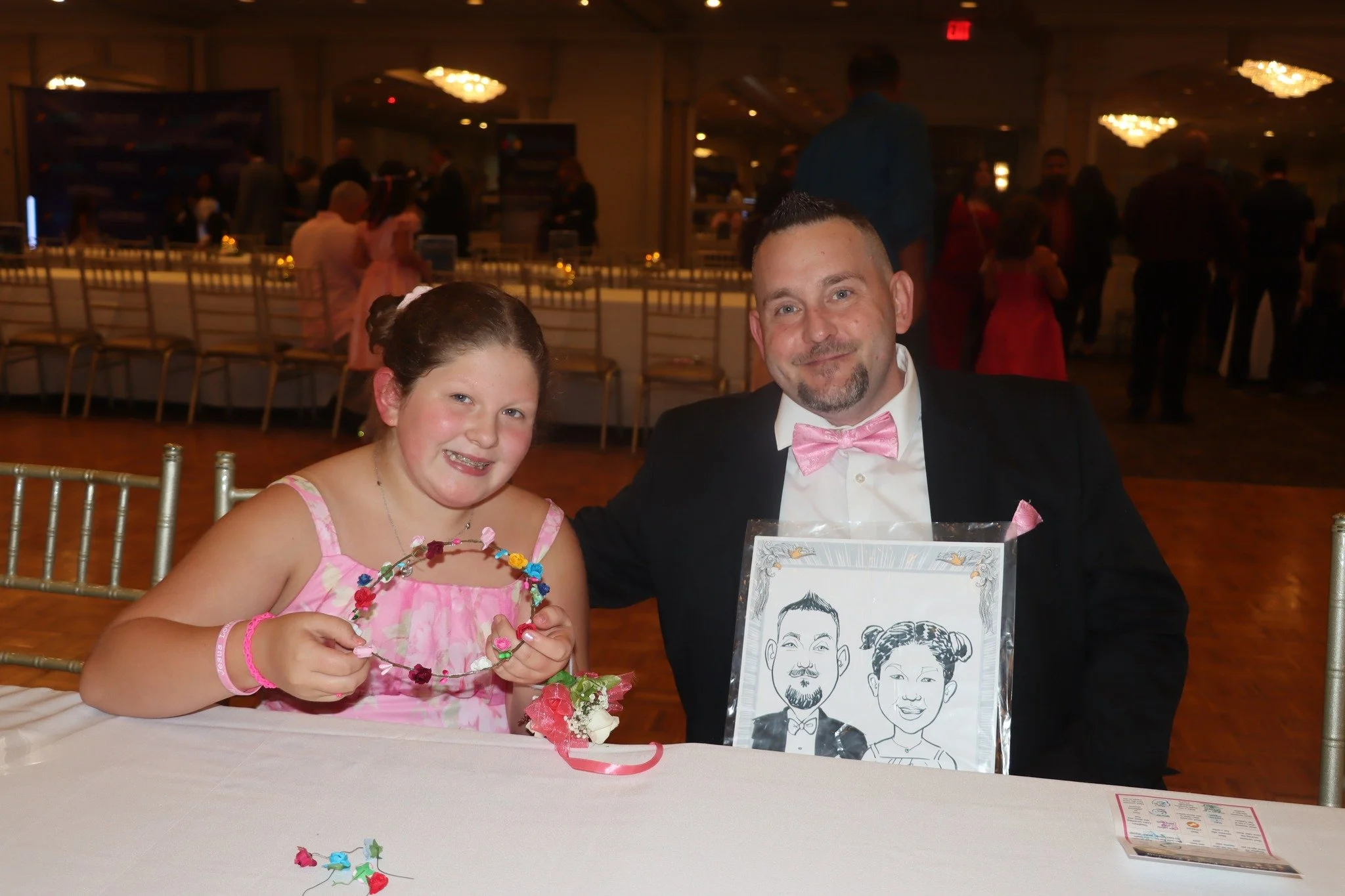 A young girl and a man in formal attire sitting at a table during a celebration. The girl is holding a colorful flower crown, and there is a caricature of a man and woman in a frame on the table. The background shows a decorated banquet hall with oth