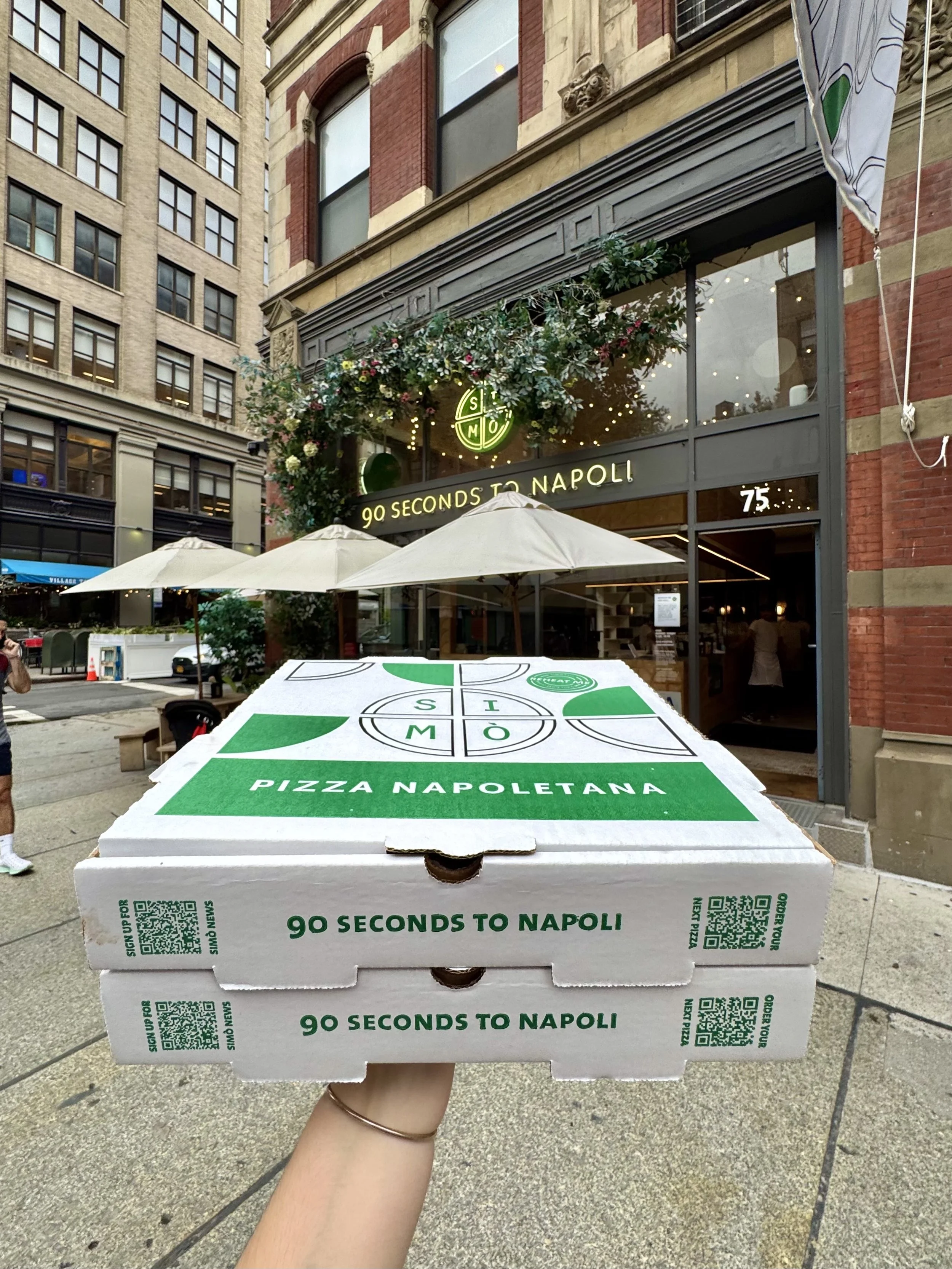 A hand holding two stacked boxes of Pizza Napoletana, with one box visible showing green and white design and the text '90 Seconds to Napoli.' In the background, there's a restaurant with signs saying '90 Seconds to Napoli,' large windows, outdoor um