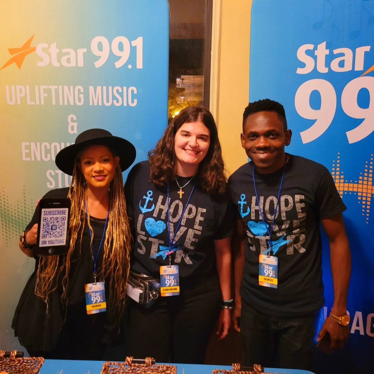 Three smiling volunteers at Star 99.1 radio station event, wearing black t-shirts with slogans 'Hope, Love, Star' and identification badges, standing in front of a promotional backdrop.
