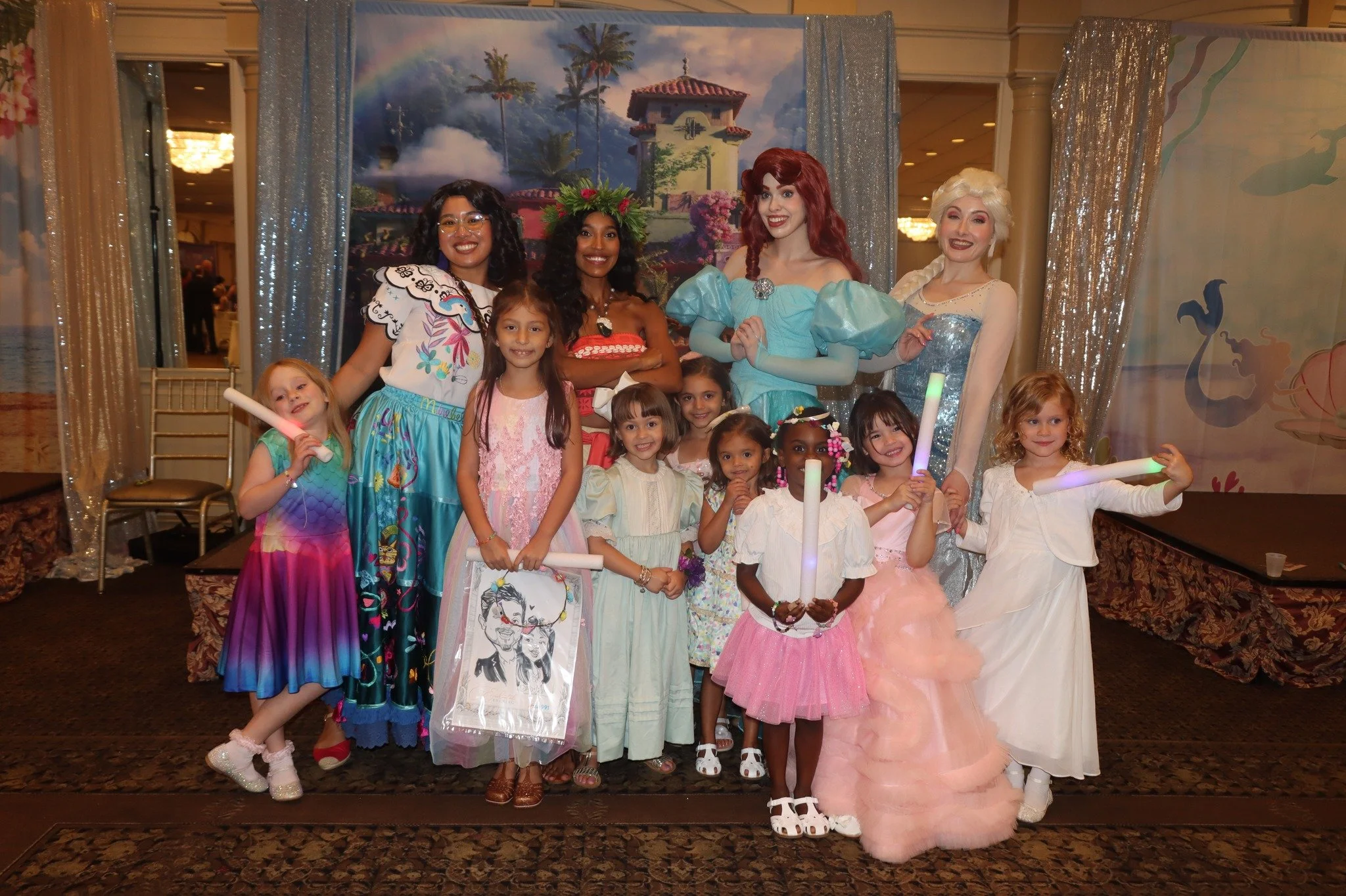 Group of young girls and women dressed as Disney princesses, posing on stage at a party. The backdrop features a scenic painting with palm trees and a rainbow. Some girls hold light-up sticks and face the camera happily.