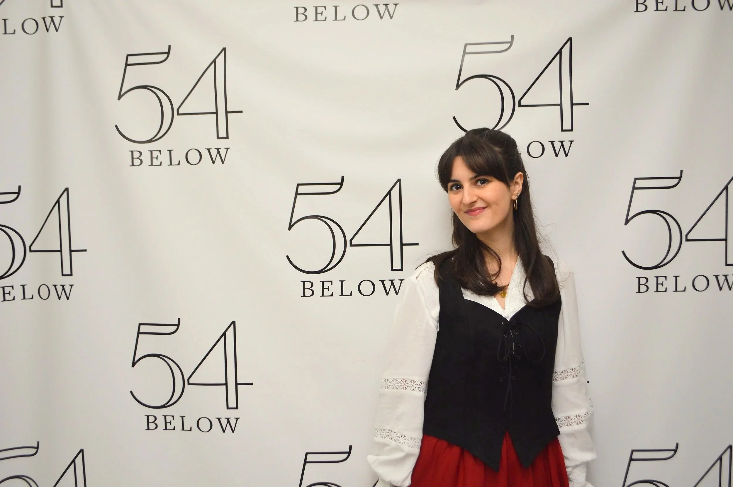 A woman with dark hair in a half-up style, wearing a white blouse with lace details on the sleeves, a black vest, and a red skirt, standing in front of a white backdrop with '54 BELOW' repeated in black text.
