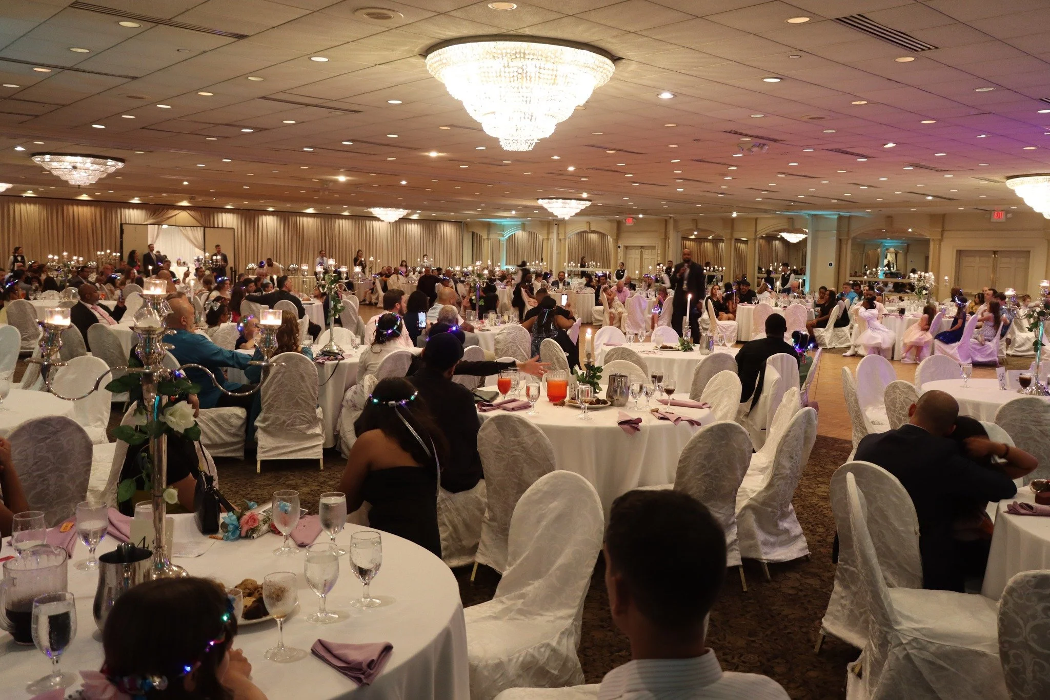 A large banquet hall decorated for a formal event with round tables, white tablecloths, flower centerpieces, and chandeliers, filled with formally dressed guests seated and socializing.