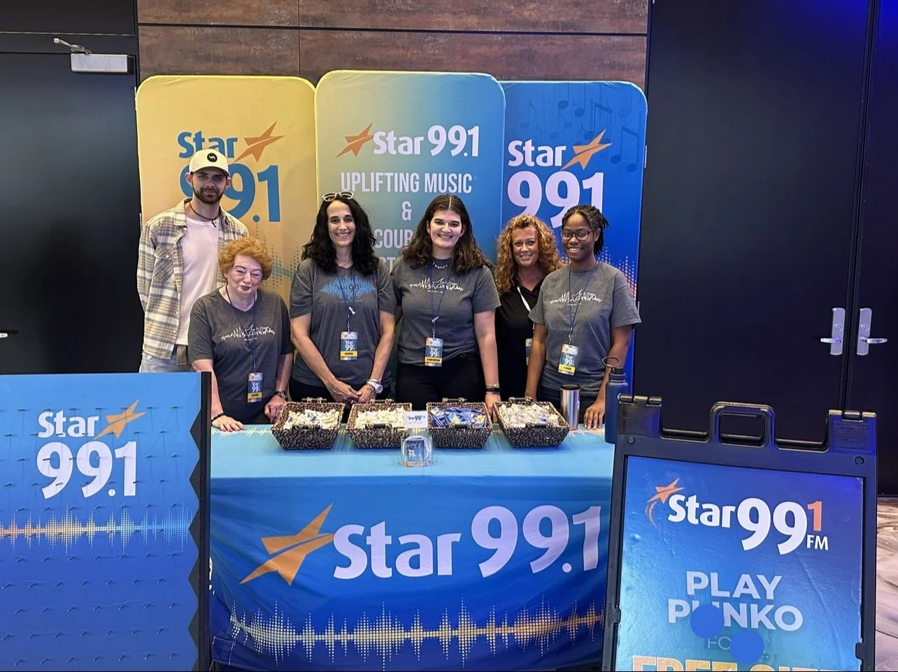 Group of six people standing behind a table with promotional materials for Star 99.1 FM radio station at an event. There are colorful banners and signs in the background advertising the station and its music focus.