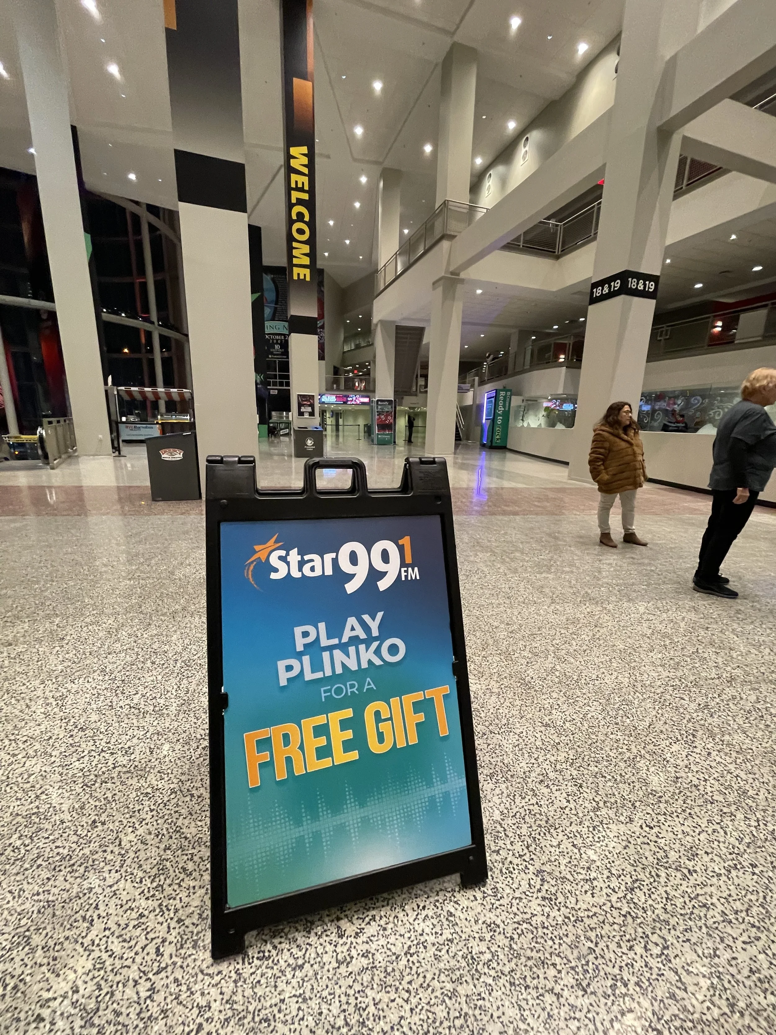 An indoor lobby of a mall or theater with a sign advertising Star 99.1 FM and a promotion to play Plinko for a free gift. The lobby has high ceilings, multiple levels, and a few people standing around.