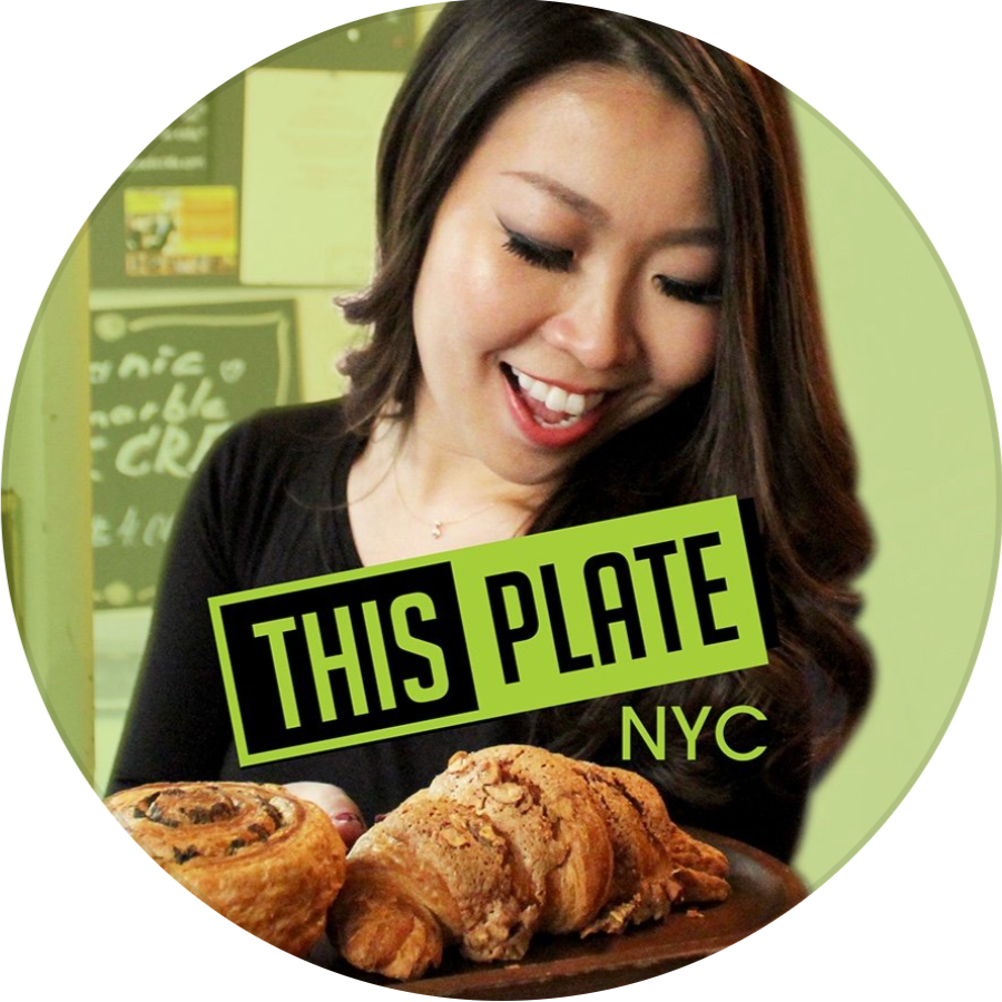 A woman with long dark hair smiling as she looks down at a plate of croissants and pastries. There is a sign in front that says "This Plate NYC." The background appears to be a cafe or bakery.