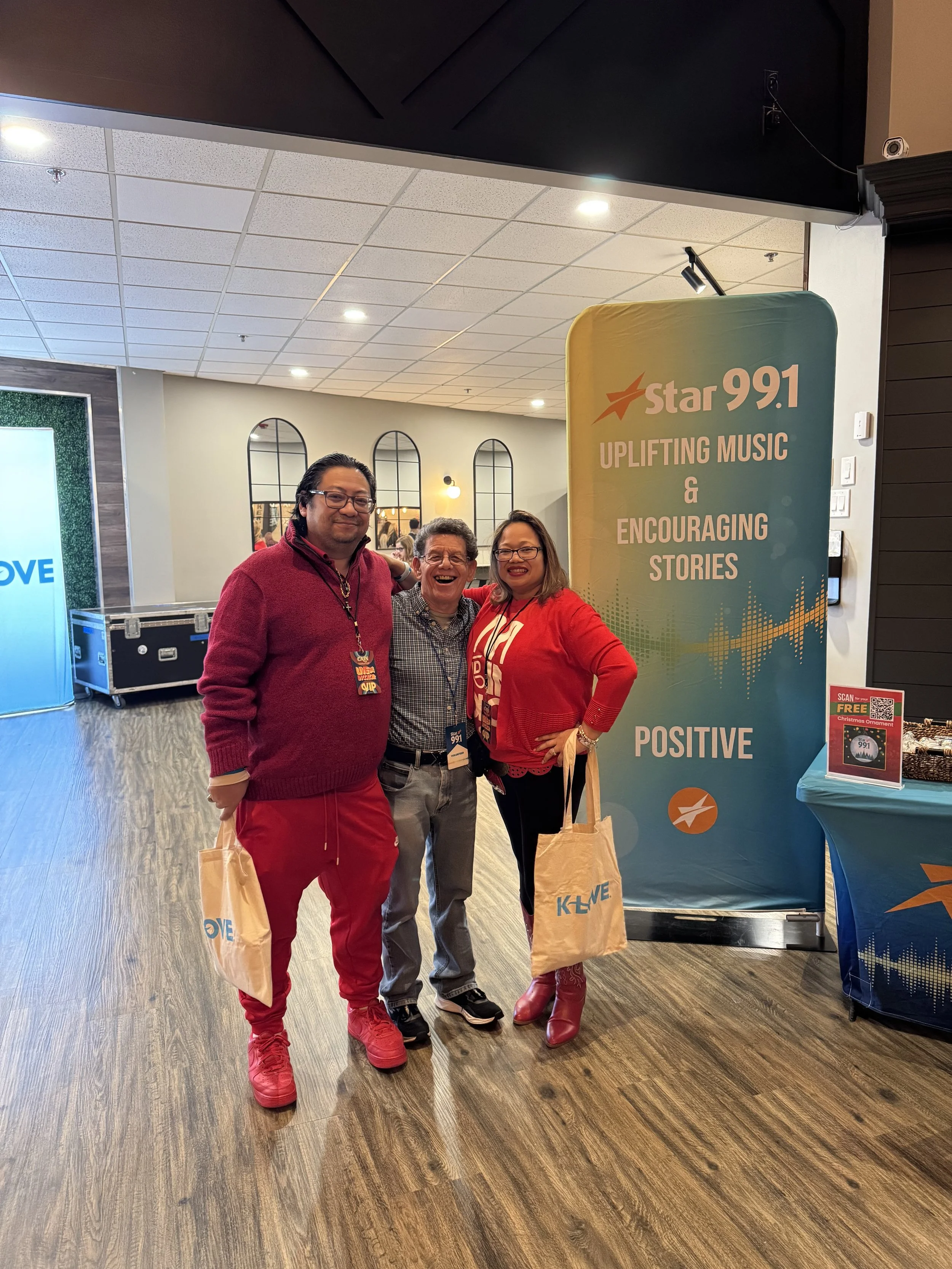 Three people smiling and posing together at an event, standing in front of a large Star 99.1 banner that promotes uplifting music, encouraging stories, and positivity.