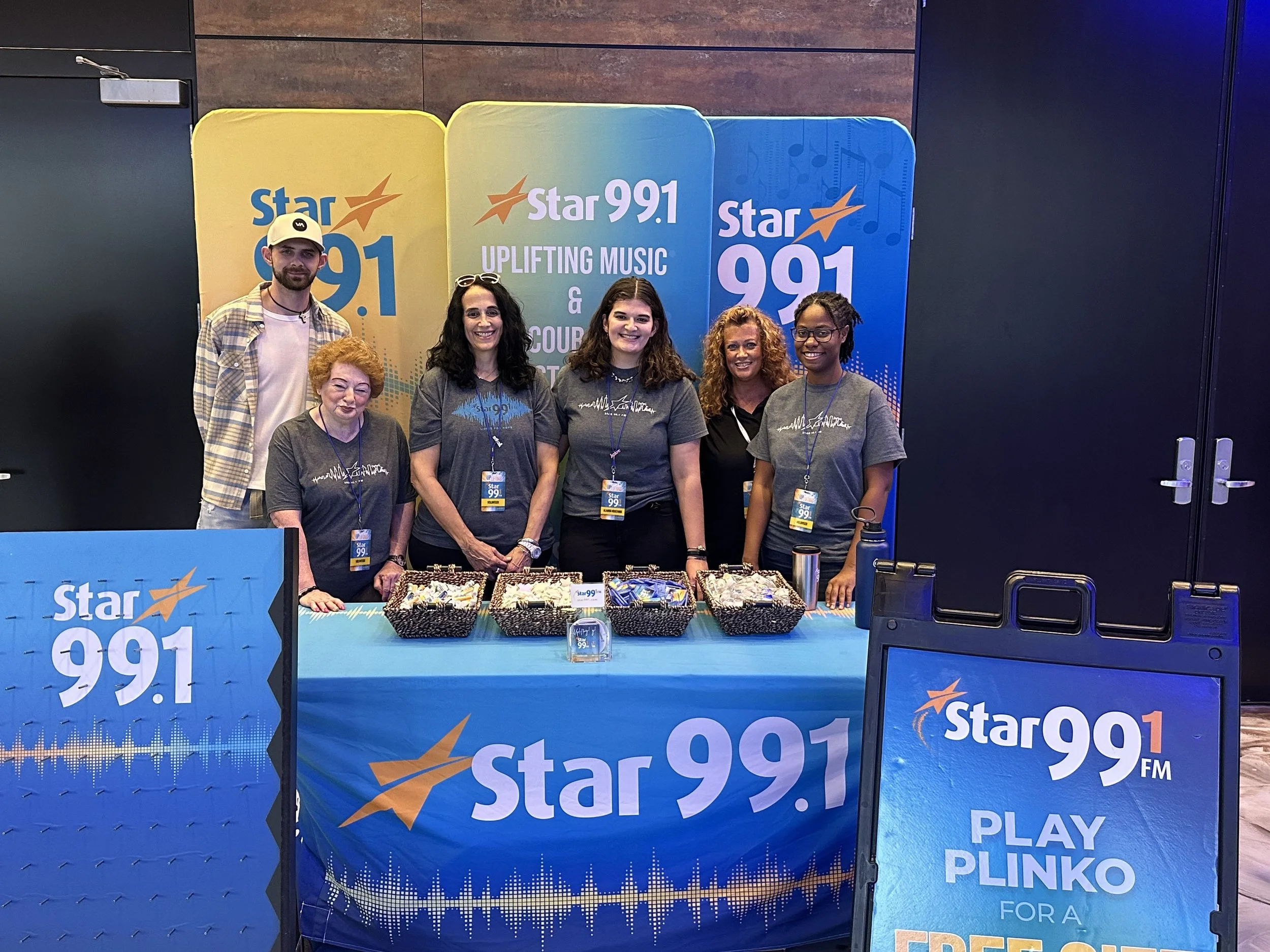 Group of six people standing behind a table at a promotional event for Star 99.1 FM, with promotional banners and snacks on the table.
