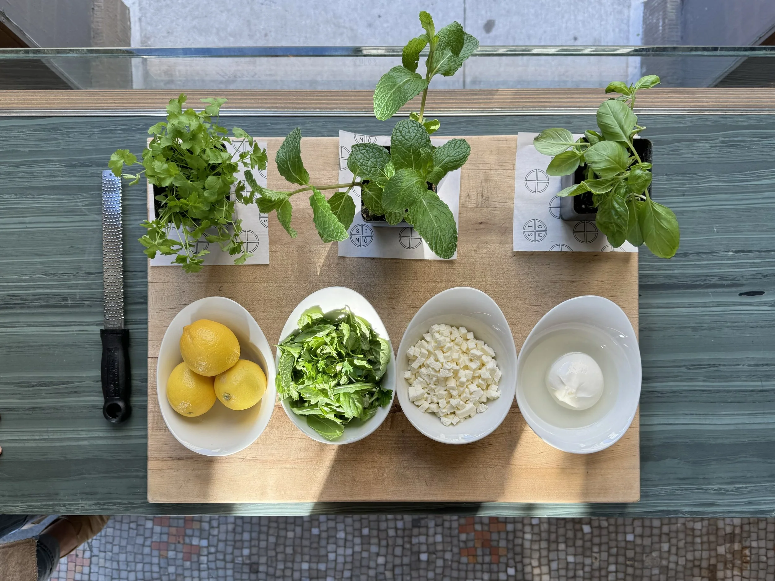 Top-down view of three potted herbs, a knife, and three bowls of ingredients on a wooden cutting board. The herbs are cilantro, mint, and basil. The bowls contain lemons, chopped cheese, and mozzarella cheese, with a bowl of milk or cream.
