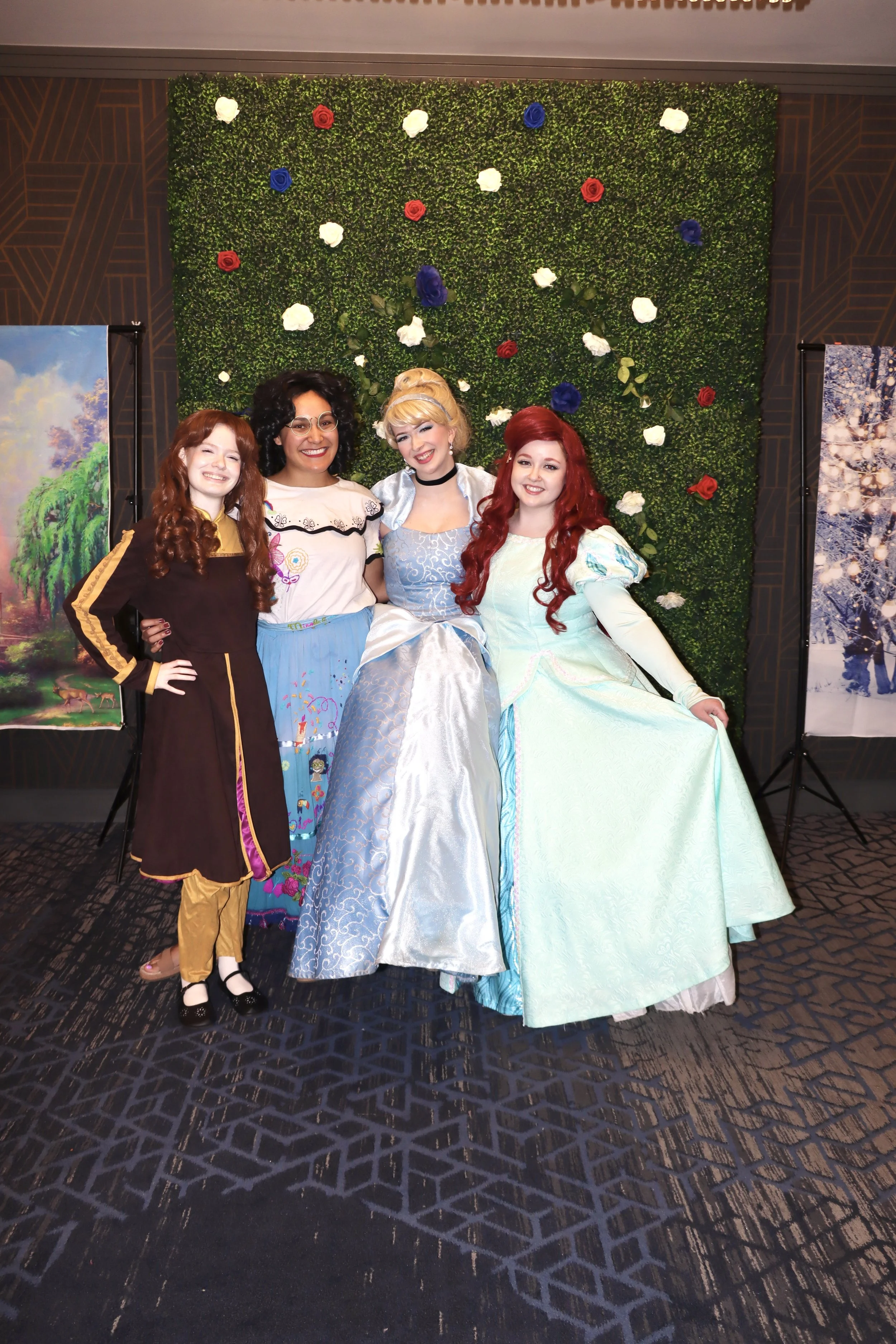 Four women dressed as characters from Disney's 'The Little Mermaid' and 'Cinderella' standing in front of a green wall decorated with white, red, and blue flowers. Two framed paintings are partially visible on either side of the group.