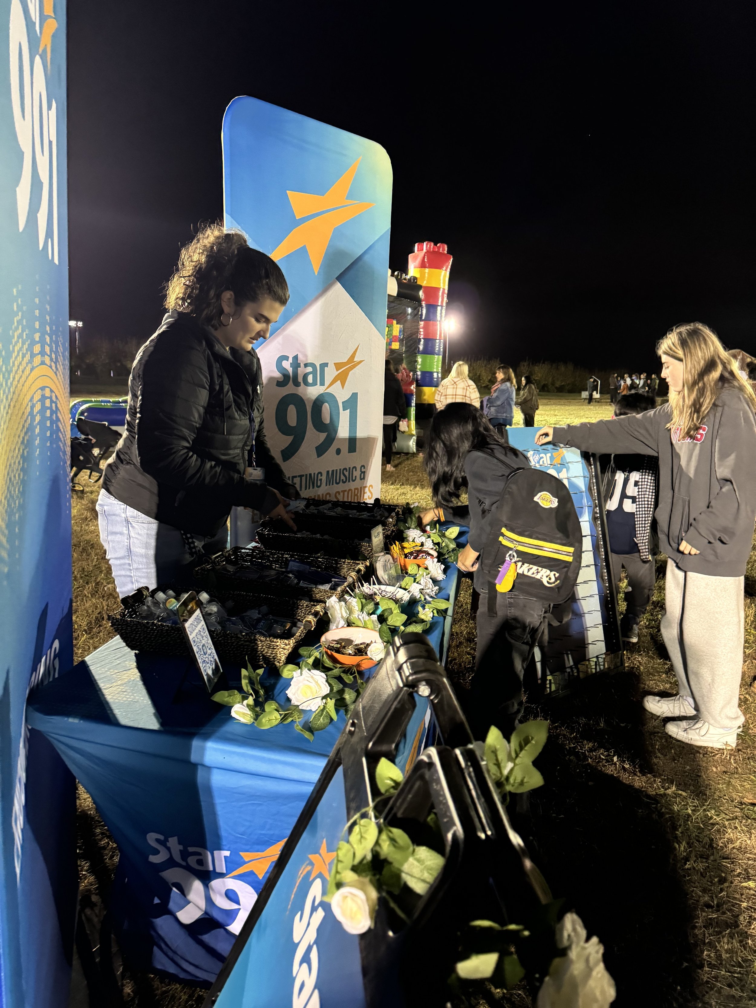 Nighttime outdoor event with a Star 99.1 radio station booth decorated with flowers and greenery, where people are browsing.