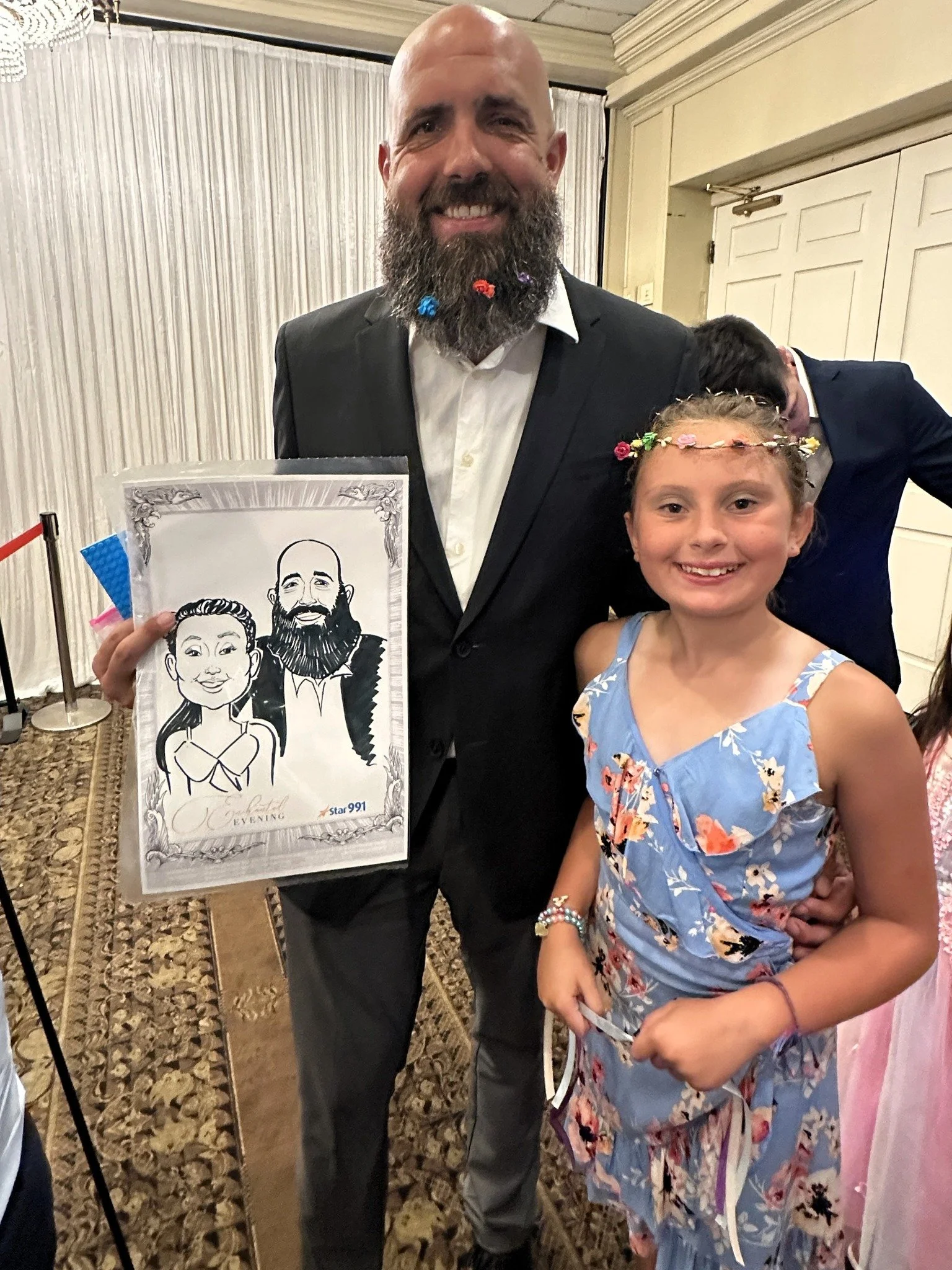 A man in a black suit holding a cartoon caricature of himself and a girl in a blue dress with a floral pattern, both smiling at a formal event.