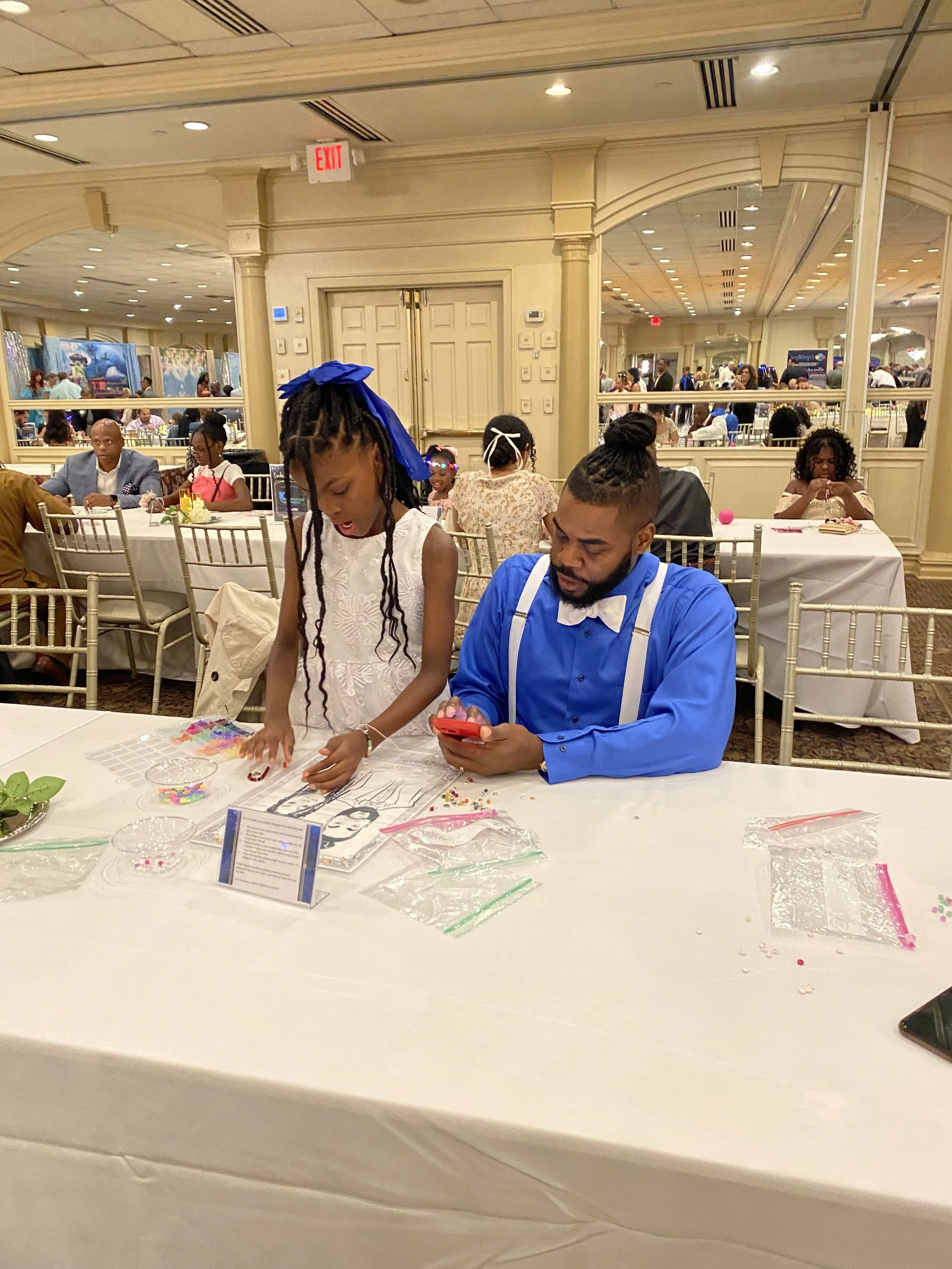 A man in a blue shirt and white suspenders sitting at a banquet table, looking at his phone, while a young girl with braided hair and a blue bow standing next to him is working on a craft project with beads and paper. The banquet hall has many guests