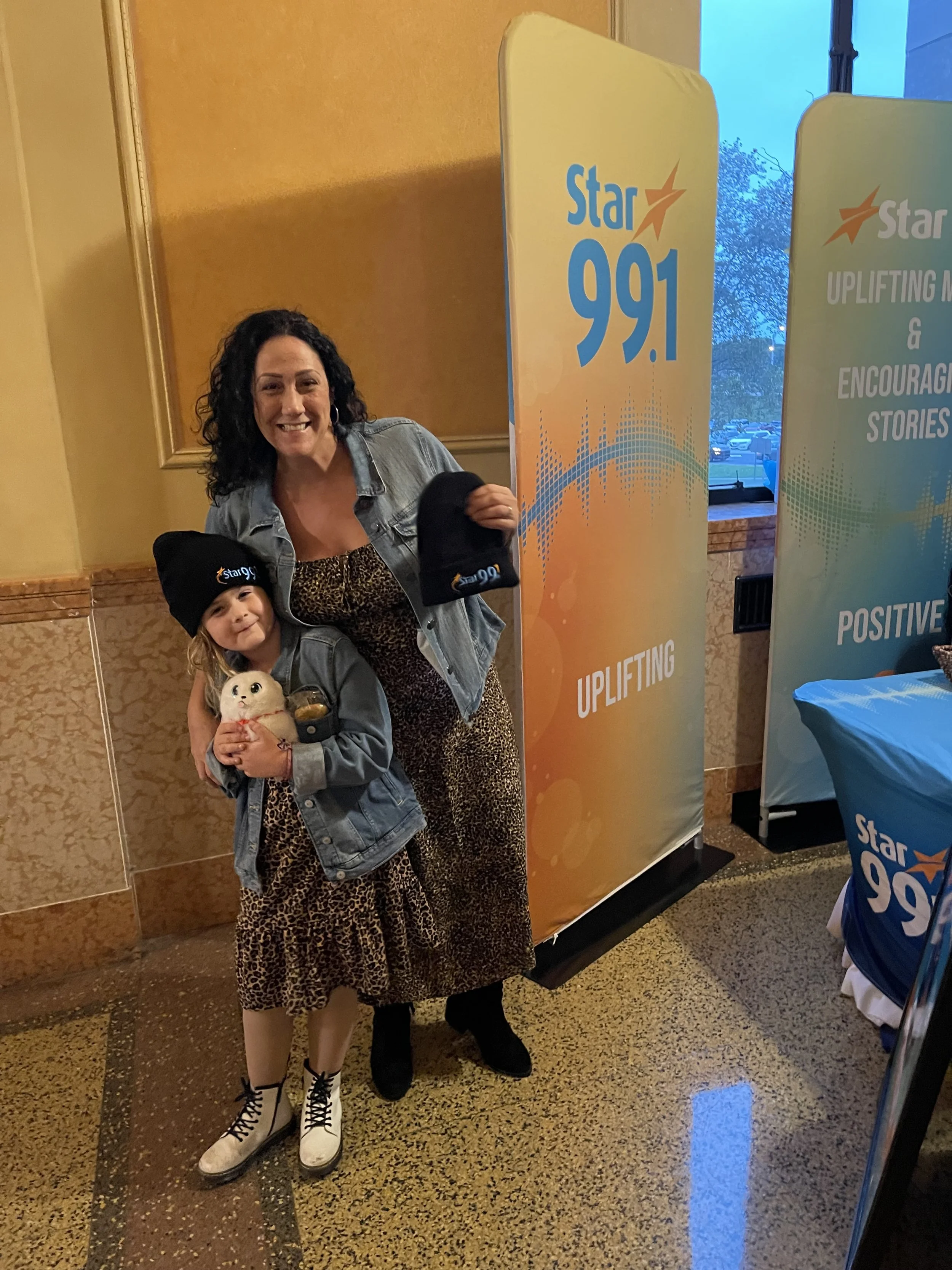 A woman and a young girl pose together indoors at a radio station event, standing next to promotional banners for Star 99.1, with the woman smiling and holding a hat, while the girl holds a small plush toy. The girl wears a beanie that says 'star 99'