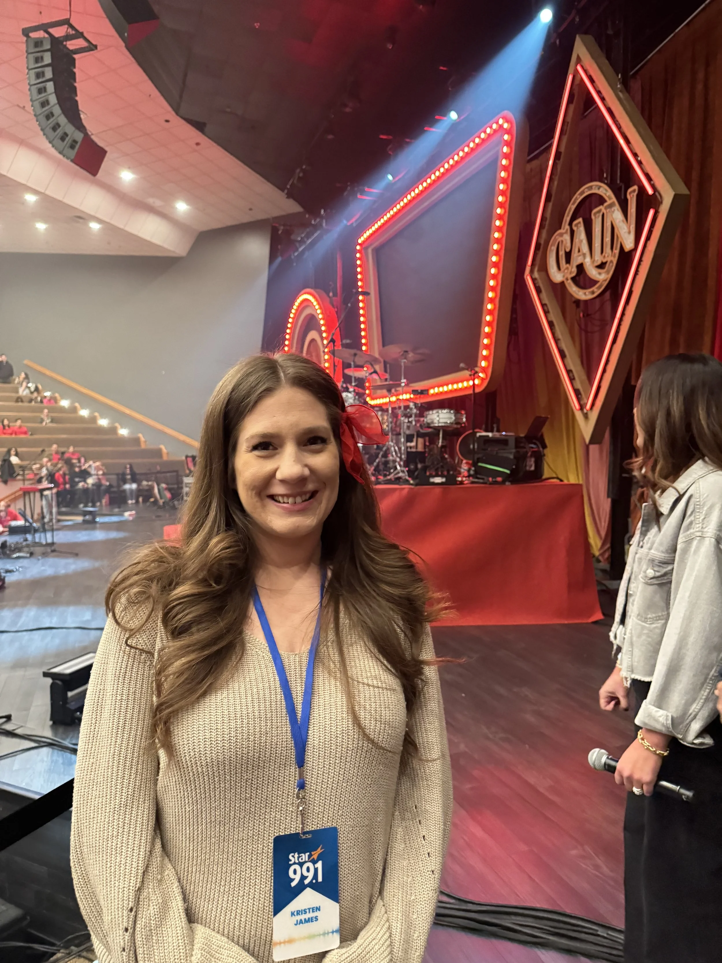 A woman with long, wavy brown hair wearing a beige sweater and a blue badge around her neck stands smiling in a theater or concert venue. In the background, a stage with musical instruments and large, illuminated red signs with the words "CAIN" and a