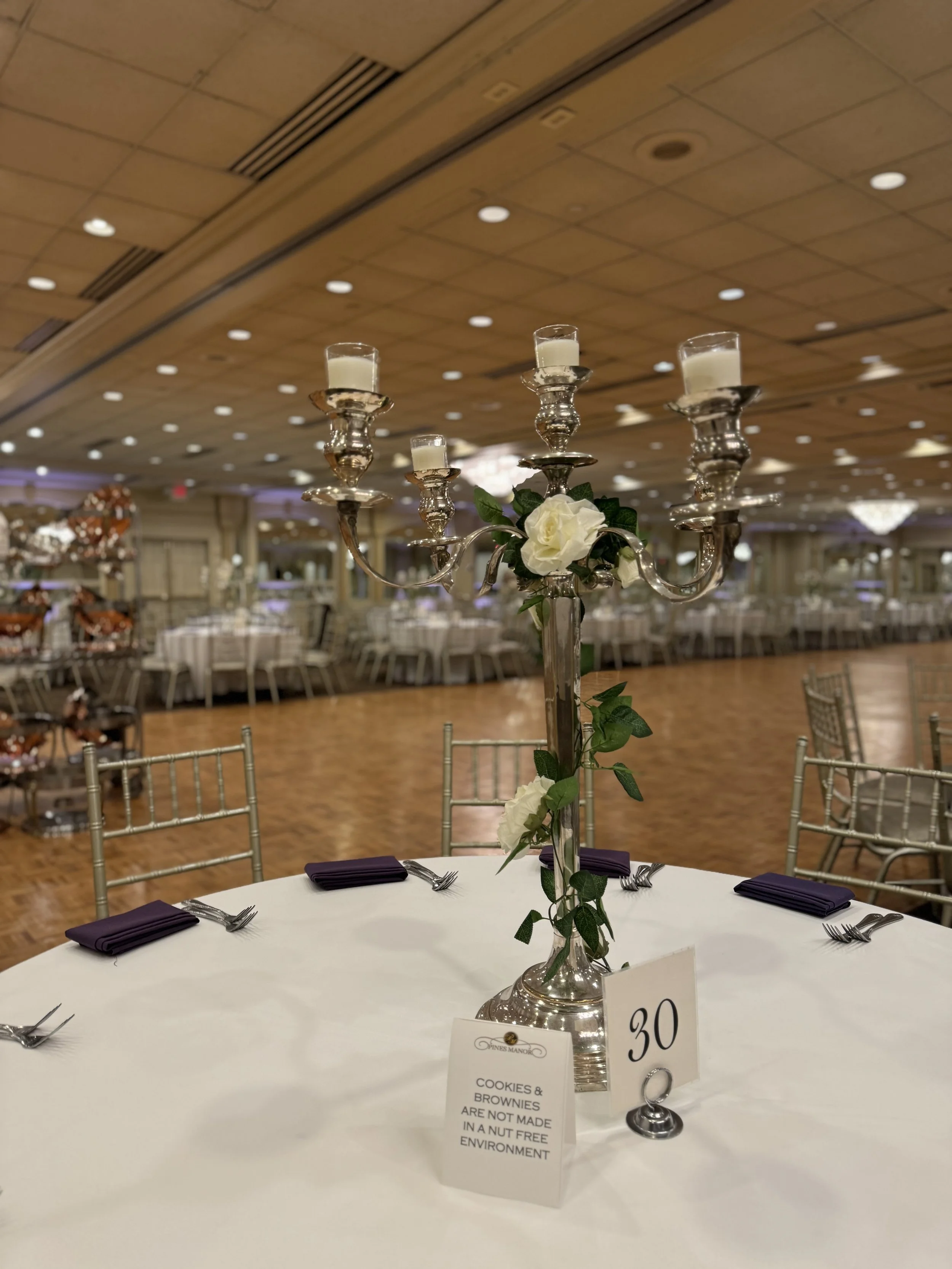 Elegant banquet table with a silver candelabrum centerpiece decorated with white roses and green leaves, set with purple napkins and silverware, in a large decorated event hall.