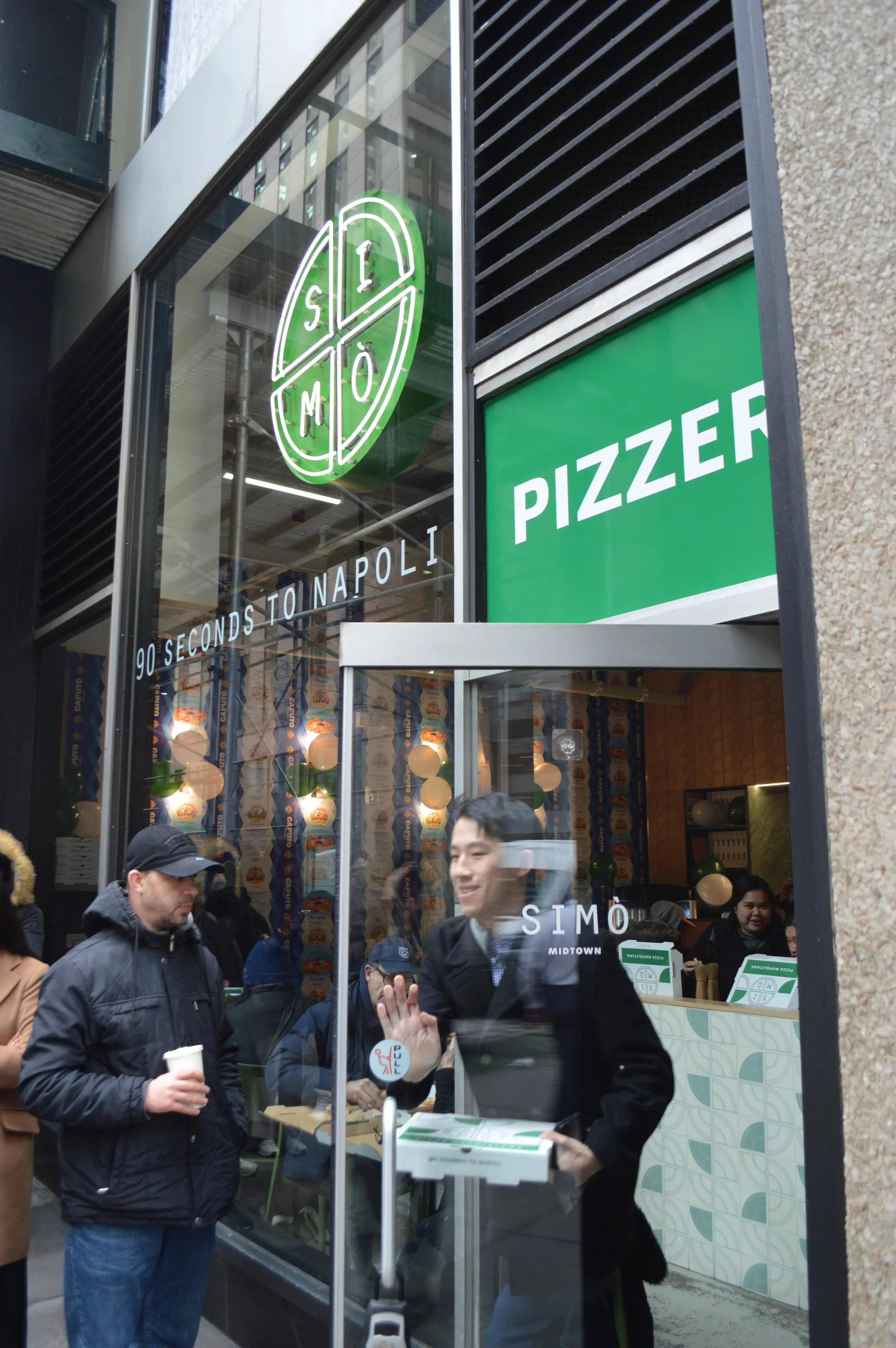 People entering a pizza restaurant named SIMÒ Midtown, with a large window displaying the logo and a sign that says 'Pizza.' Inside, customers are seated and staff are visible behind the counter.