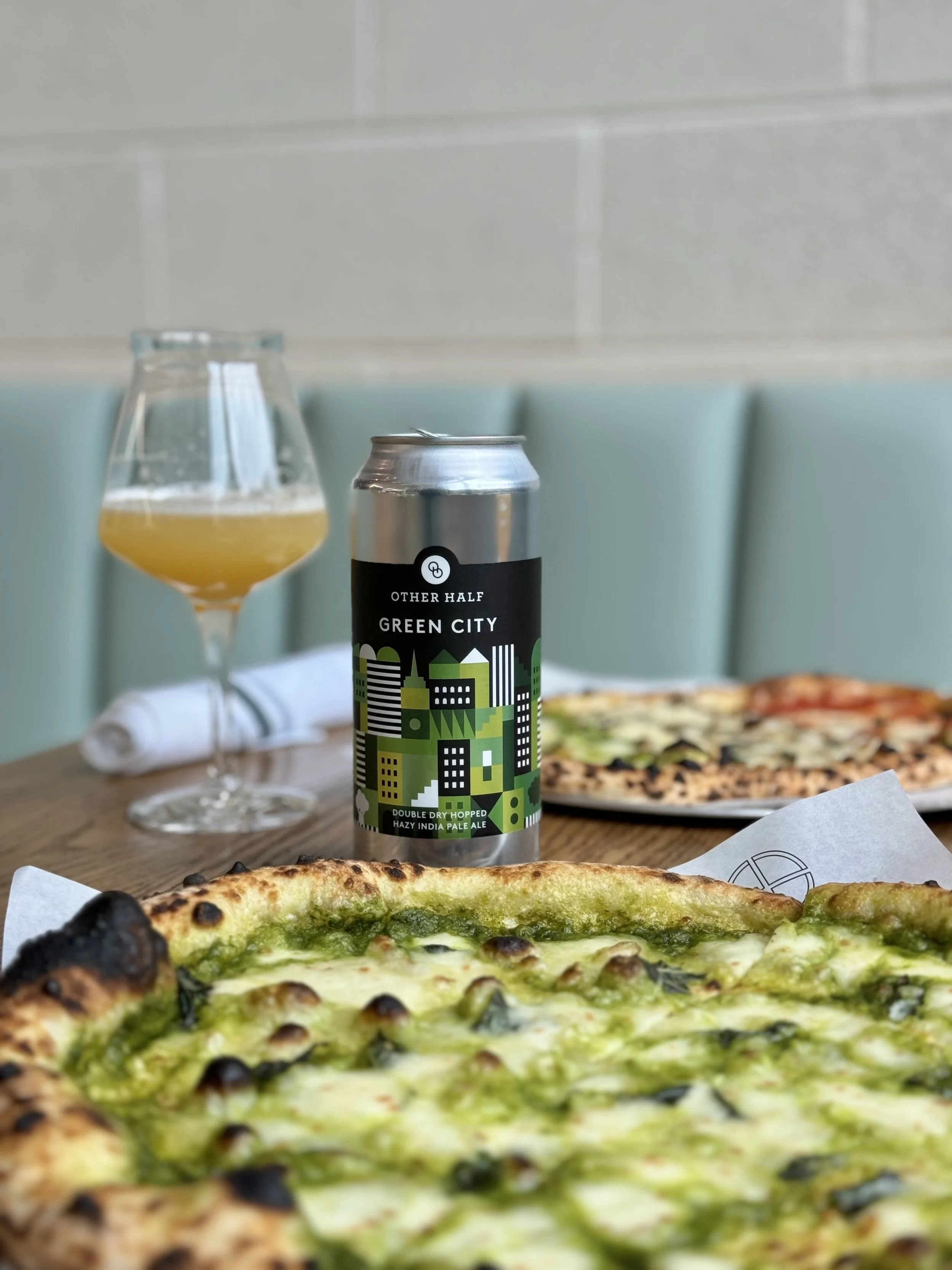 Pizza with green pesto and toppings, a can of Green City India Pale Ale, and a glass of beer on a wooden table in a restaurant setting.