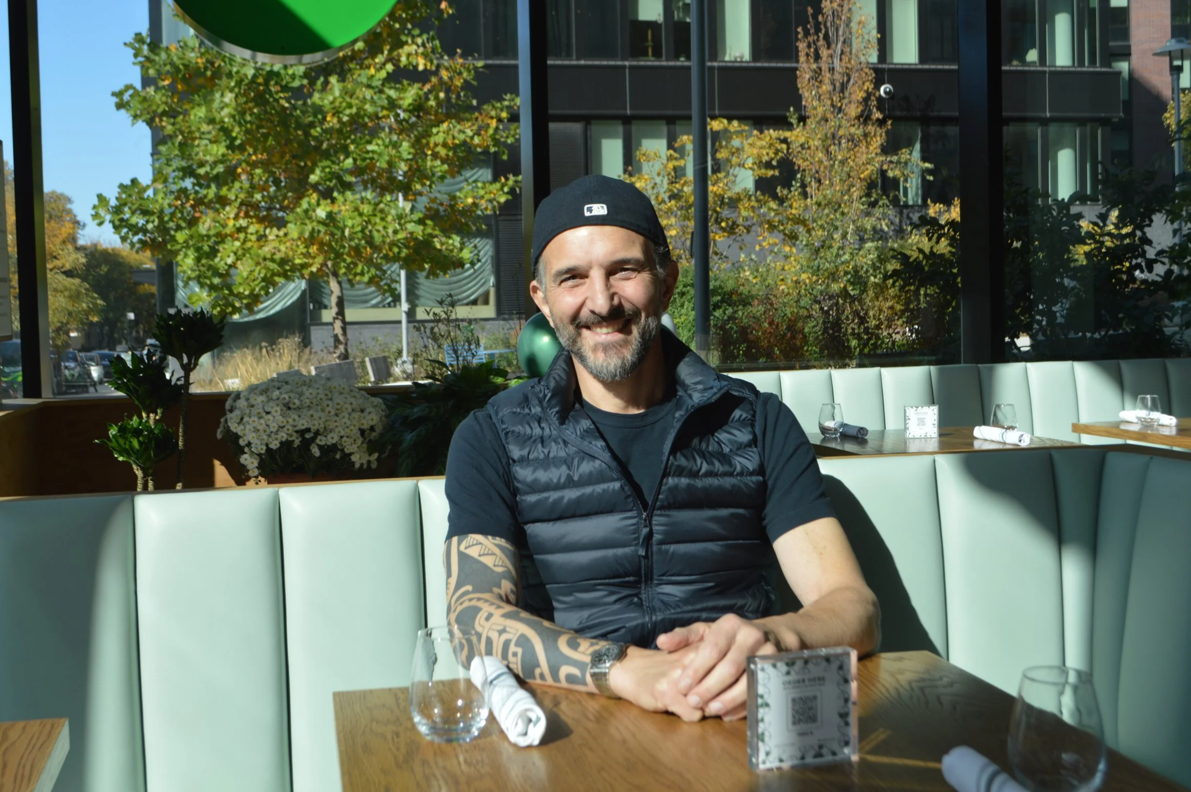 Smiling man with tattoos sitting at a restaurant table with glassware, napkin, and a QR code, in a modern restaurant with large windows and outdoor trees.