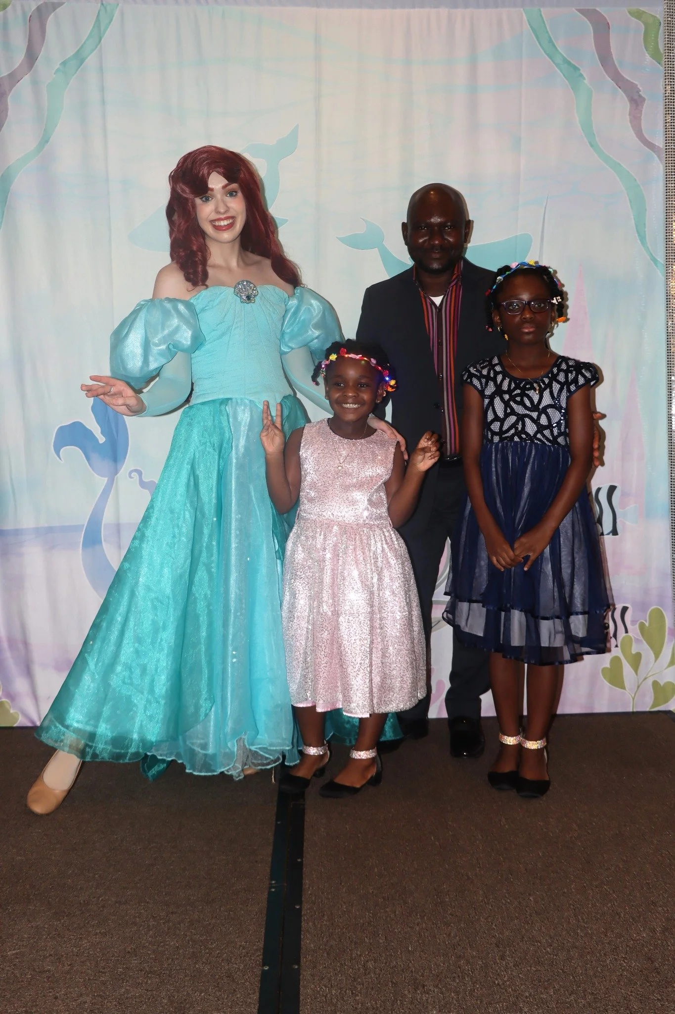 A woman dressed as Ariel from Disney's The Little Mermaid poses with a man and two young girls in front of a colorful backdrop featuring mermaid and seaweed designs.